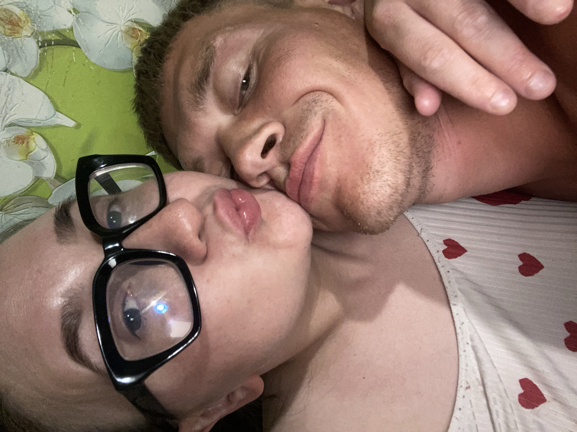 A close-up shot shows two people lying down. The man on the right has short blond hair and stubble. He is smiling slightly, with his eyes open and looking at the camera. His hand is partially visible at the top right corner of the image. The woman on the left wears black-framed glasses, and her lips are pursed as if she is about to kiss someone. A bright reflection is visible in the lens of her glasses. The background is composed of green floral patterned sheets on the left and a white shirt with red hearts on the right. There is no information to confirm that this image was taken in the city of Chaikovsky, Russia.
