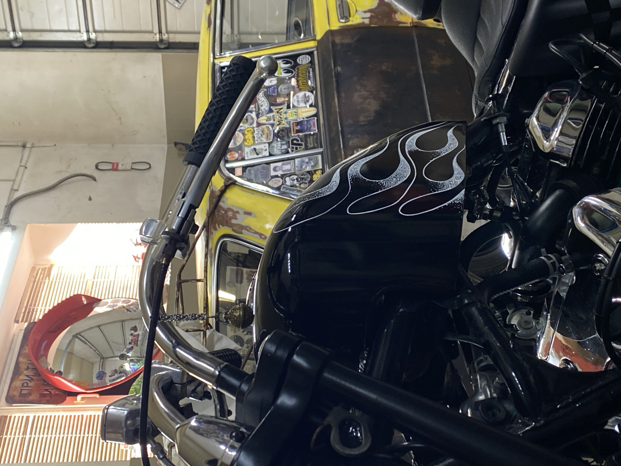 The image depicts a close-up view of a black motorcycle, foregrounded by its handlebars and fuel tank which features a white flame decal. Behind the motorcycle, a vintage yellow car is visible, decorated with numerous stickers on its rear window. The car appears to be parked in a garage or workshop, evidenced by a roll-up garage door in the background and fluorescent lighting. A red convex mirror is mounted above the motorcycle's handlebars, reflecting a portion of the garage interior. A sign with Cyrillic text is partially visible on the wall behind the mirror. The overall scene suggests a custom motorcycle and car enthusiast's space. The location is identified as Agronom, Russia.