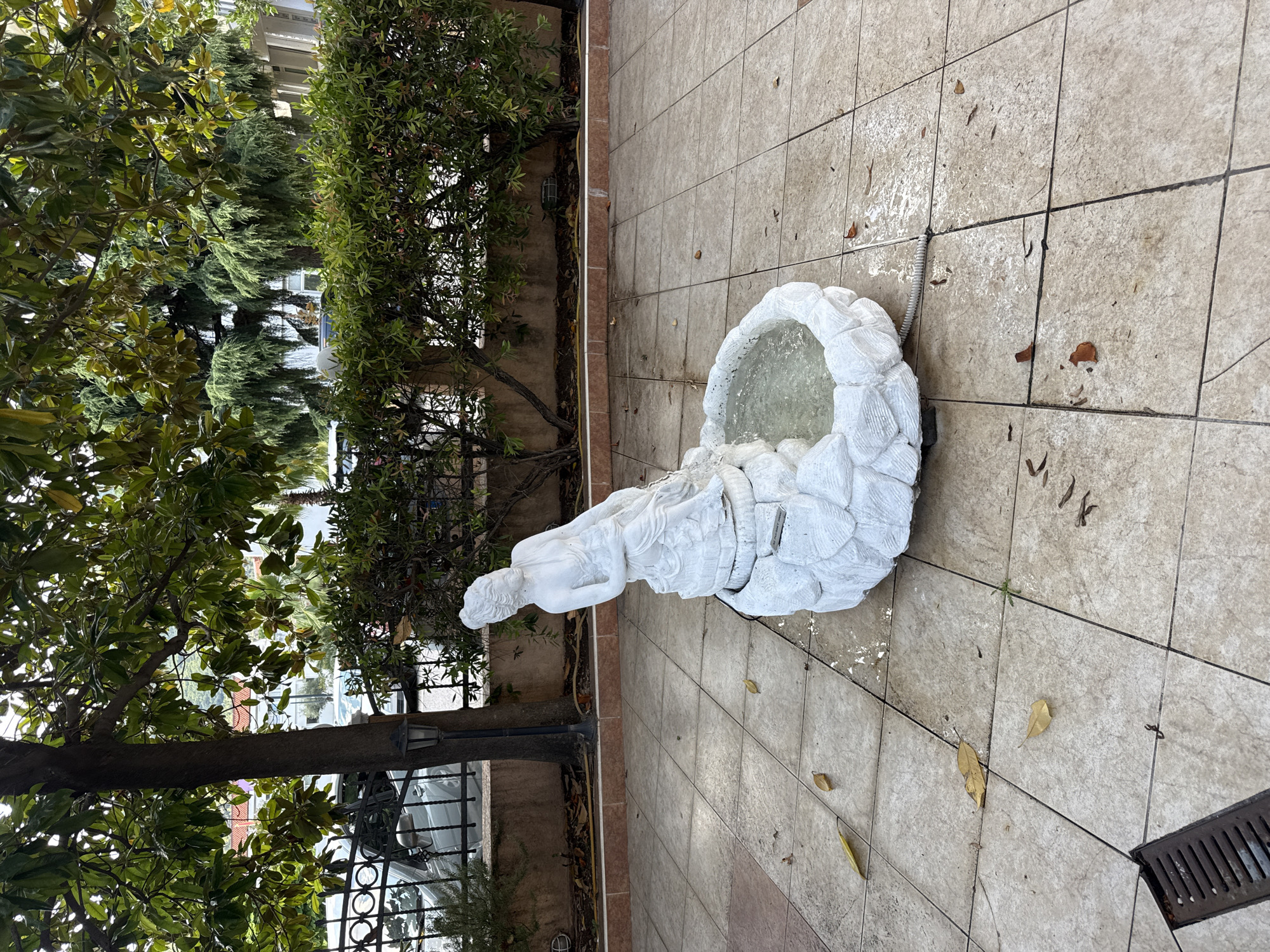 The image shows a white statue of a woman fountain, with water filling its basin. The statue is located outdoors on a tiled patio. Green foliage from trees and bushes surrounds the patio. The time of day appears to be daytime, with natural light illuminating the scene. The weather seems to be mild, possibly after a rain shower, as the tiles are wet and some leaves are scattered on the ground. The overall setting is that of a garden or courtyard, likely in a Mediterranean climate given the mention of Budva, Montenegro. The statue itself depicts a reclining woman whose body forms part of the fountain's structure. The water is clear and reflects the surrounding elements. There is a drain grate visible on the right side of the image. The composition suggests a peaceful and tranquil atmosphere.