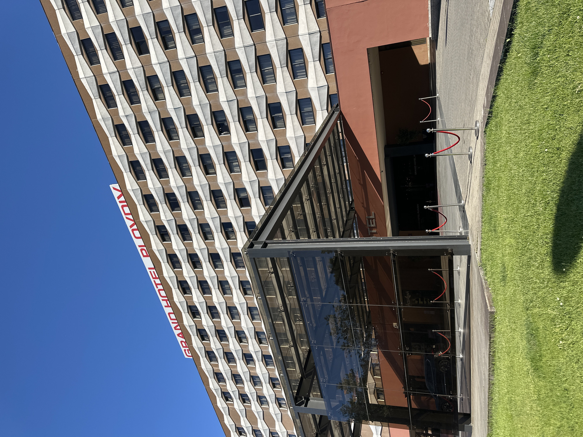The image shows the Grand Hotel Plovdiv in Plovdiv, Bulgaria. The hotel is a tall building with a distinctive facade of alternating brown and white geometric shapes, each containing a dark rectangular window. The name "GRAND HOTEL PLOVDIV" is vertically displayed in red lettering on the side of the building. The hotel's entrance features a modern glass and metal canopy. Red velvet ropes on silver stanchions guide foot traffic toward the entrance. Adjacent to the building is a sidewalk bordered by a patch of green grass. The sky is a clear, bright blue.