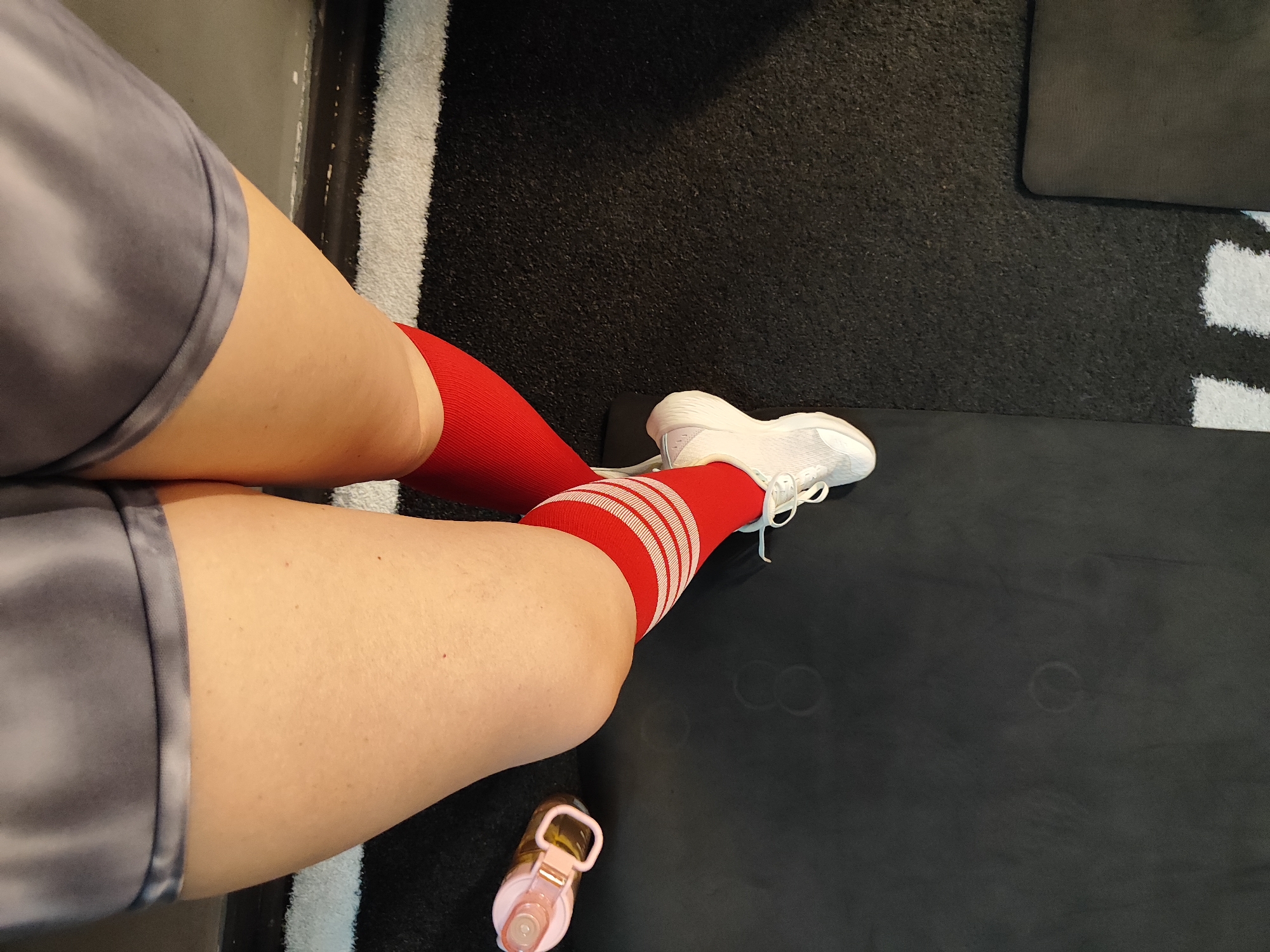 The image shows a close-up view of a person's legs wearing red athletic socks with white stripes and grey shorts. They are also wearing white athletic shoes. A pink water bottle is visible near their feet. The person is seated on a black mat, which appears to be in a gym or fitness studio. The background consists of a textured black surface and a white striped border. There is no discernible information in the image to indicate a specific location in Almaty, Kazakhstan.
