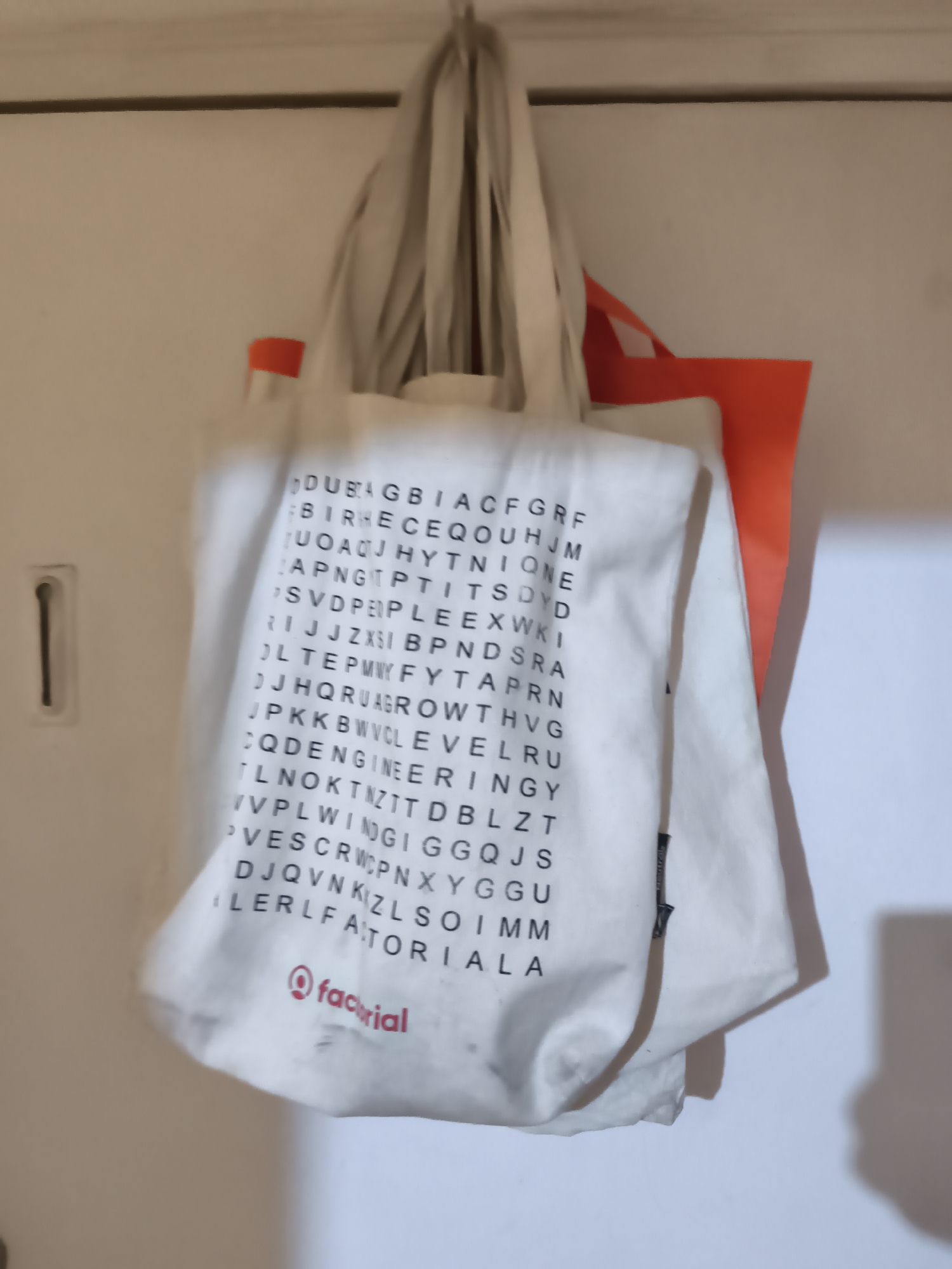 A close-up shot shows two tote bags hanging on what appears to be a wall or a door. The bag in the front is white with black lettering arranged in a grid, resembling a word search puzzle. The word "FACTORIAL" is prominently displayed in a red logo near the bottom of the bag. Behind the white bag is an orange tote bag, partially visible. The bags seem to be hanging from hooks. The environment appears to be indoors, possibly a home or office space.