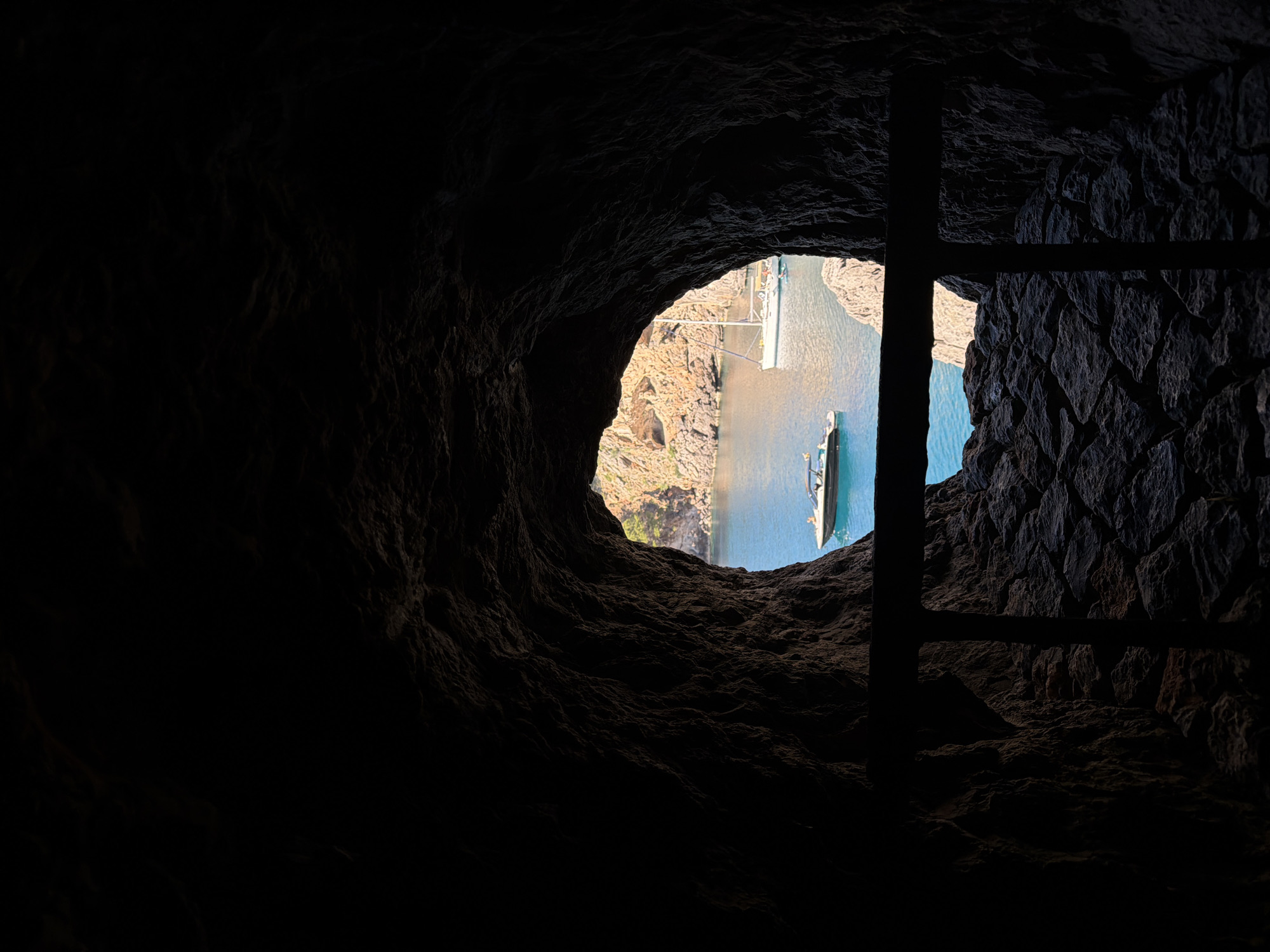 A dimly lit cave in Escorca, Spain, opens to a view of water and rocky cliffs, punctuated by a distant boat. The cave walls are rough-hewn, stone, and an indistinct black structure, possibly a ladder, partially obscures the opening.