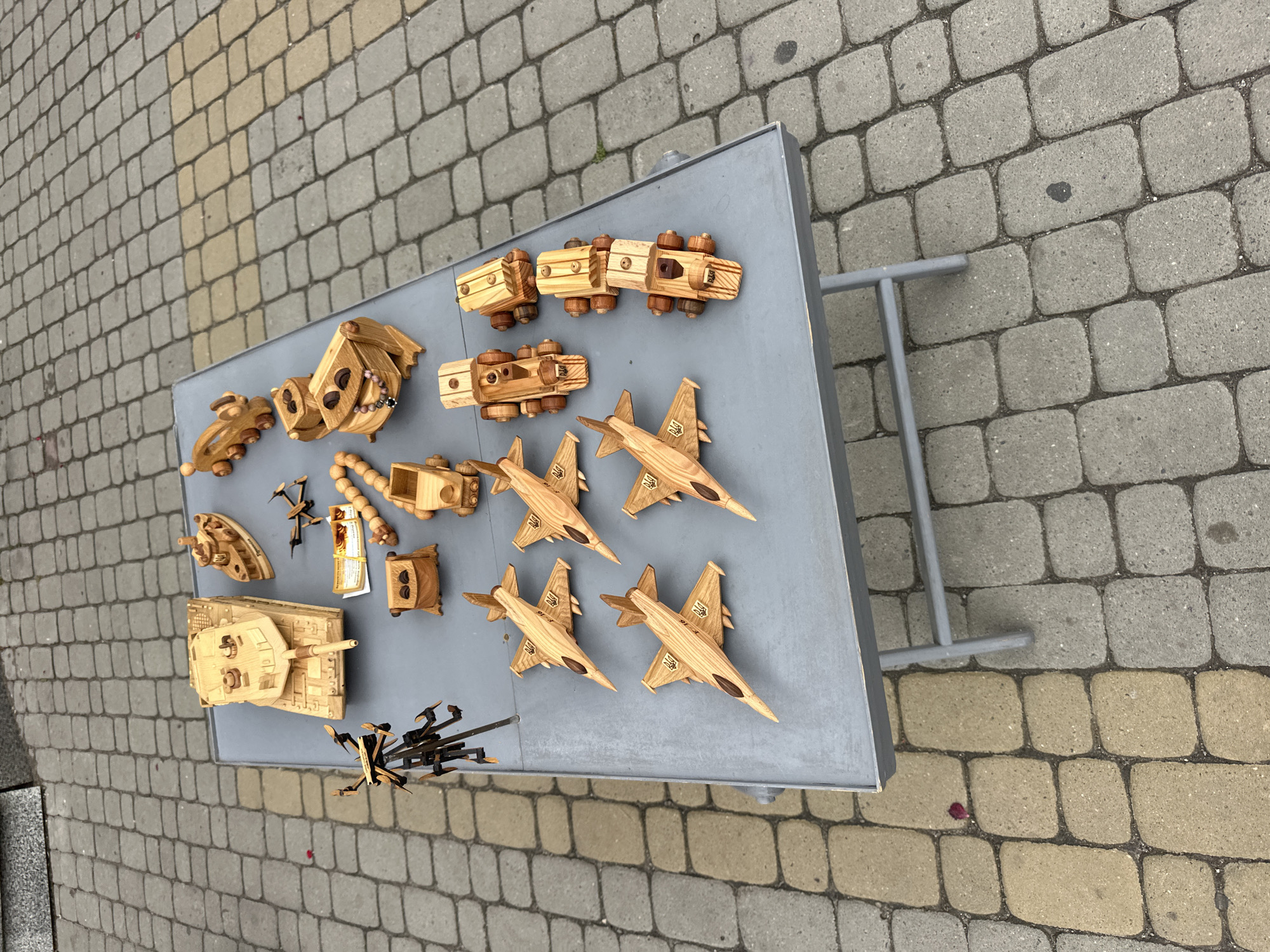The image shows a display of wooden toys on a grey table, likely at a street market or fair in Ternopil, Ukraine. The toys are intricately carved and include a variety of vehicles such as tanks, airplanes, and trains, as well as some abstract figures. The table is placed on a cobblestone pavement, typical of many European cities. The background suggests an outdoor setting, with the paving stones extending out of frame. There are no people visible in the immediate vicinity of the table, though the context implies a public space where people would be browsing. The lighting is natural, indicating it is daytime. The overall impression is one of handmade crafts and a local artisan's work.