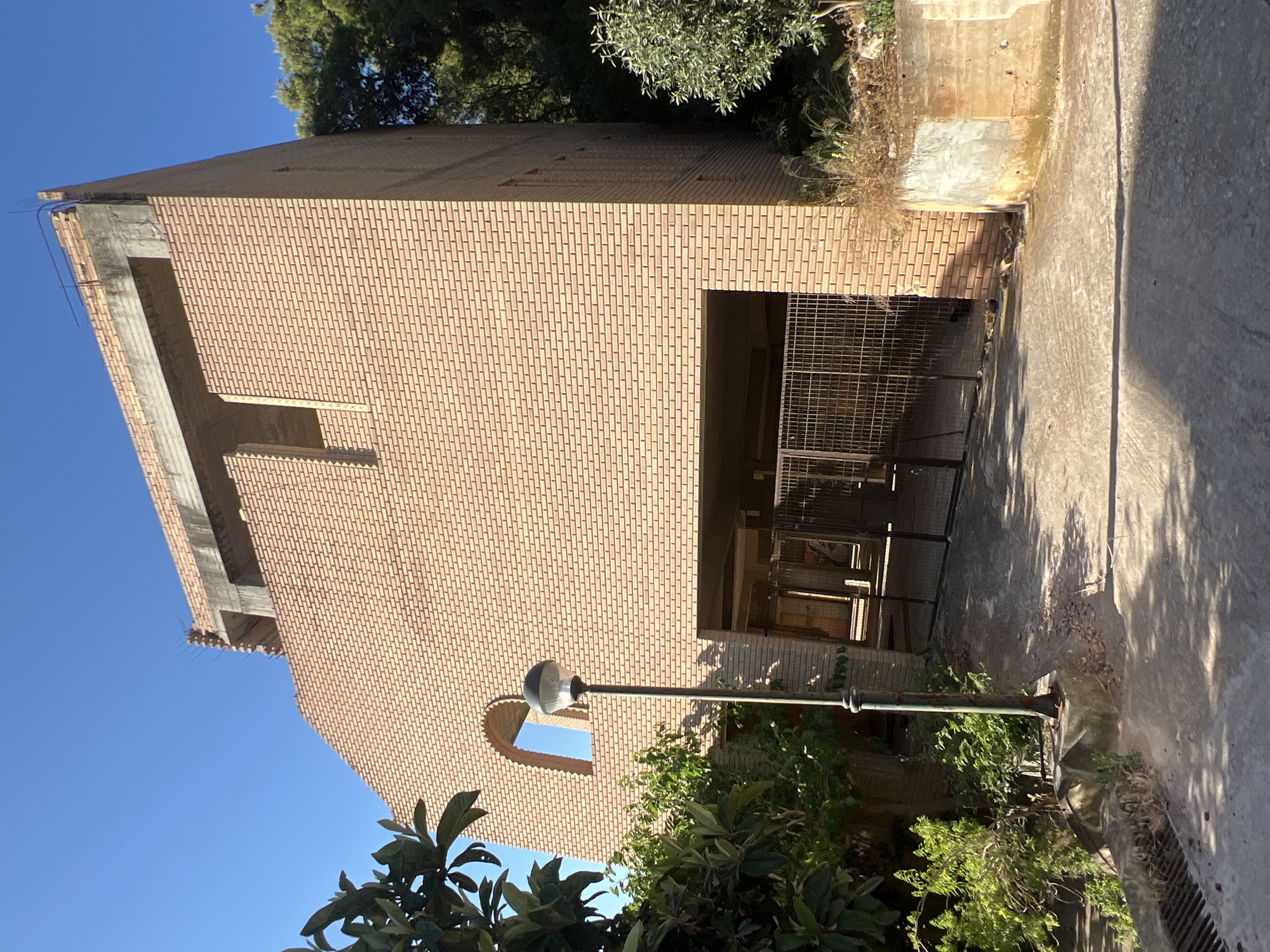 The image depicts an unfinished brick building with exposed rebar and concrete at the top, suggesting construction is ongoing or has been halted. The building is situated in an outdoor environment with a clear blue sky. To the left of the building, there are trees and lush green vegetation. A single lamppost stands in front of the building, and a driveway leads to a large opening with a metal gate, which appears to be an entrance to a garage or parking area. The ground is paved with concrete. The scene is located on a sunny day, casting shadows on the ground. The provided crops confirm the architectural details and surrounding vegetation.