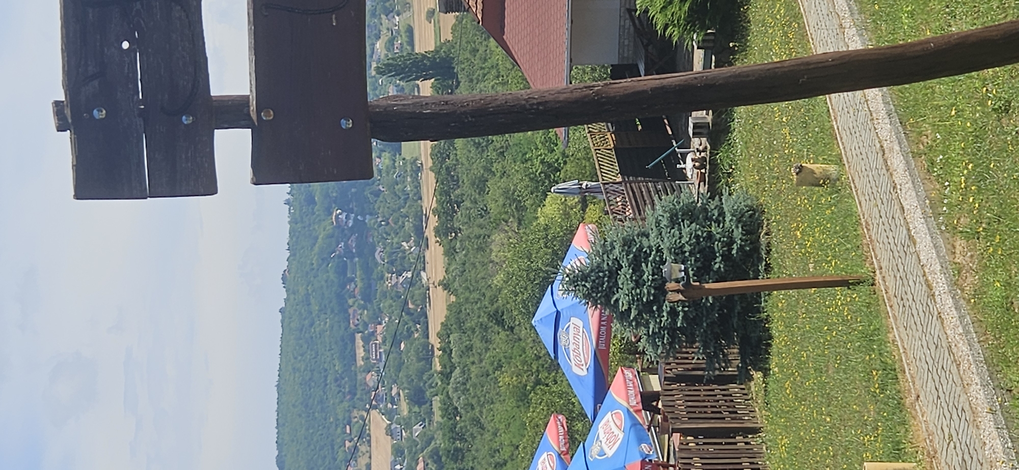 The image depicts a scenic view from an elevated position in Hévíz, Hungary. In the foreground, a weathered wooden post with two dark wooden signs is positioned to the right, partially obscuring the view. A paved walkway runs diagonally across the bottom of the frame, bordered by grassy areas dotted with yellow flowers. To the left of the walkway, a small evergreen tree stands near a wooden fence. Beyond the fence, several blue and red outdoor umbrellas, advertising "Kobanyai," are set up, suggesting an outdoor seating area for a restaurant or cafe.
In the mid-ground, a village with scattered houses is nestled amongst lush green trees and fields. The architecture appears to be typical of Hungarian rural settlements. A more distant mountain range, possibly the Keszthely Mountains, is visible on the horizon, with a light haze obscuring its finer details. The sky is partly cloudy with a bright, hazy atmosphere, indicating a warm, sunny day. A small building with a red-tiled roof is visible to the right, and a tall, slender tree stands out against the village backdrop. A small bird is perched on a yellow post to the right of the walkway.