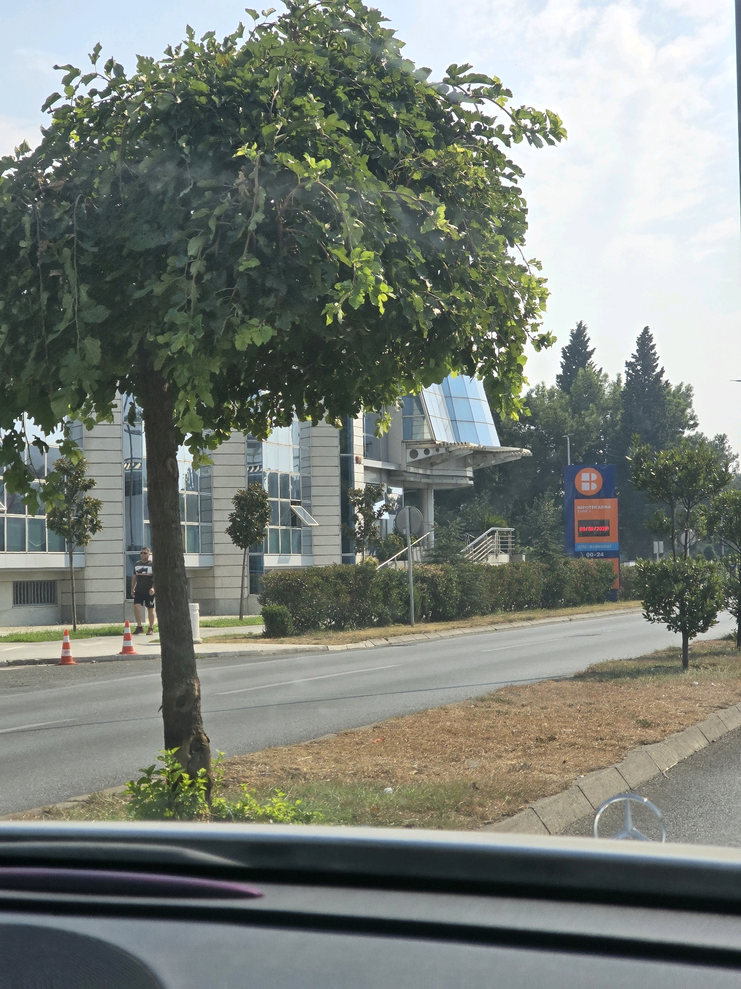 The image shows a street view in Podgorica, Montenegro, with a focus on a modern office building and surrounding greenery. In the foreground, the dashboard of a car is visible, with the Mercedes-Benz logo reflected. A mature tree with a dense canopy of green leaves stands prominently in the mid-ground, partially obscuring the building. To the right of the tree, a man in a t-shirt and shorts is walking on the sidewalk. The building behind him is a multi-story structure with a facade of light-colored panels and large, reflective windows, some of which are open. A contemporary architectural element, possibly a cantilevered section with glass panels, extends from the building.
Further to the right, a tall, orange and blue vertical sign displays the logo of Hipotekarna Banka. Text on the sign indicates "ATM - BANKOMAT 00-24," suggesting it's a 24-hour ATM service. Several coniferous trees and smaller leafy trees are interspersed around the building and along the roadside. The street in front of the building is paved asphalt, with a dry, grassy verge and a concrete curb. Two orange traffic cones are placed on the sidewalk near the tree. The sky is bright with scattered clouds. The overall scene suggests an urban environment during daylight hours.