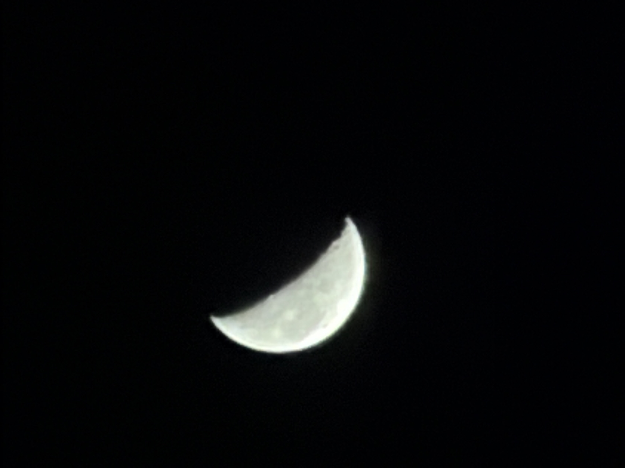 A crescent moon is visible in a dark sky. The moon is positioned in the lower left quadrant of the image. Its illuminated portion is a bright white, while the unilluminated portion blends into the blackness of space. The moon's surface shows some texture, suggesting craters or other geological features. The background is entirely black, indicating a night scene. There is no information within the image that suggests a specific location in Kandersteg, Switzerland.
