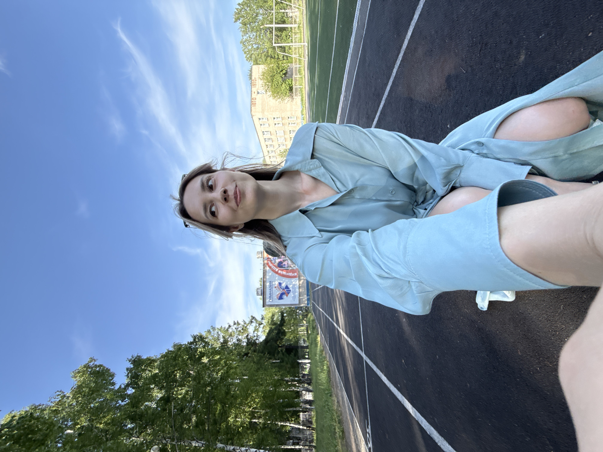 A person, likely female, sits on a running track under a partly cloudy blue sky. The track is marked with white lines and is bordered by grass and trees. A building with "1870-1910" inscribed on it is visible in the background. A billboard with Cyrillic text and logos stands near the track. The photograph appears to be taken in Chaikovsky, Russia, based on the Cyrillic text on the billboard. The person is wearing a light blue shirt or dress.