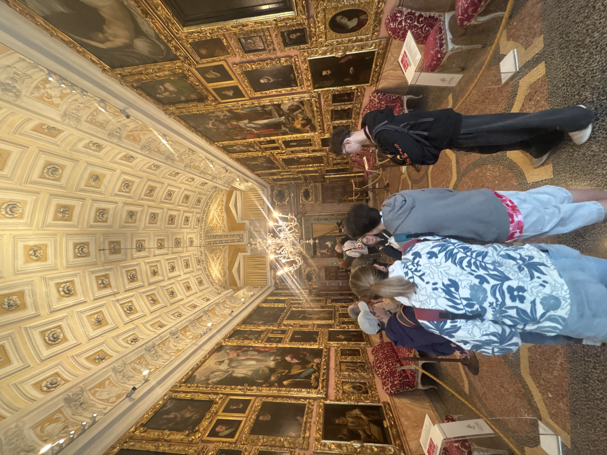The image captures a grand, ornate interior hall, likely an art gallery or palace, from a tilted perspective. The long, narrow space features a richly detailed, coffered ceiling, predominantly light yellow and white, decorated with sculptural rosettes and intricate molding. A large, multi-tiered crystal chandelier hangs from the ceiling, casting bright light into the room.
The walls are densely adorned with numerous framed oil paintings, stacked from floor to ceiling. These artworks, housed in elaborate gilded frames, appear to be classical portraits and historical or religious scenes. Below the paintings, along the walls, are sections of red, patterned velvet benches or seating. The floor is a mosaic or patterned stone, exhibiting geometric designs in earthy tones of brown, beige, and reddish-brown.
Several people, appearing to be visitors or tourists, are present in the hall, mostly seen from behind or in partial profile. They are observing the artworks and the grand architecture. One person in the foreground wears a white and blue floral patterned shirt. Another wears a black hoodie and backpack, looking upwards. Small white informational signs or brochures are visible on stands near the floor. The scene depicts a quiet, contemplative atmosphere typical of a museum visit within a historically significant building in Carciano, Italy. No visible text on the signs is legible.