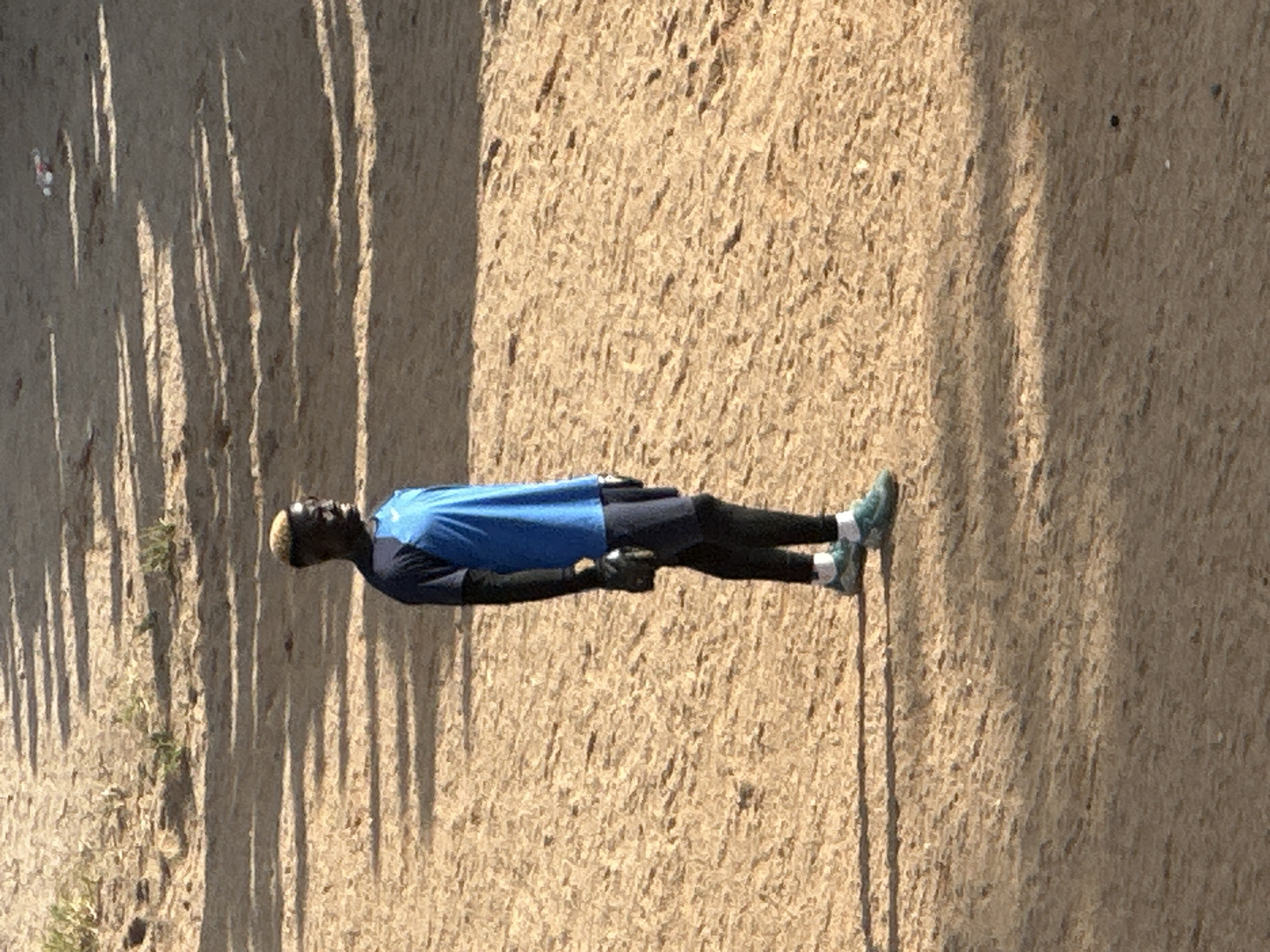 A person stands on a sandy surface in what appears to be a sports field, possibly in Jos, Nigeria, given the context. The individual is wearing a blue and black athletic top, black leggings, and teal athletic shoes. They are facing to the right of the frame, with their body angled slightly away from the viewer. The ground is covered in sand and dirt, with shadows cast by what appears to be foliage or a structure from the left side of the image. There are some scattered green plants visible in the upper left corner of the frame. The overall lighting suggests it is daytime, with the sun casting distinct shadows.
