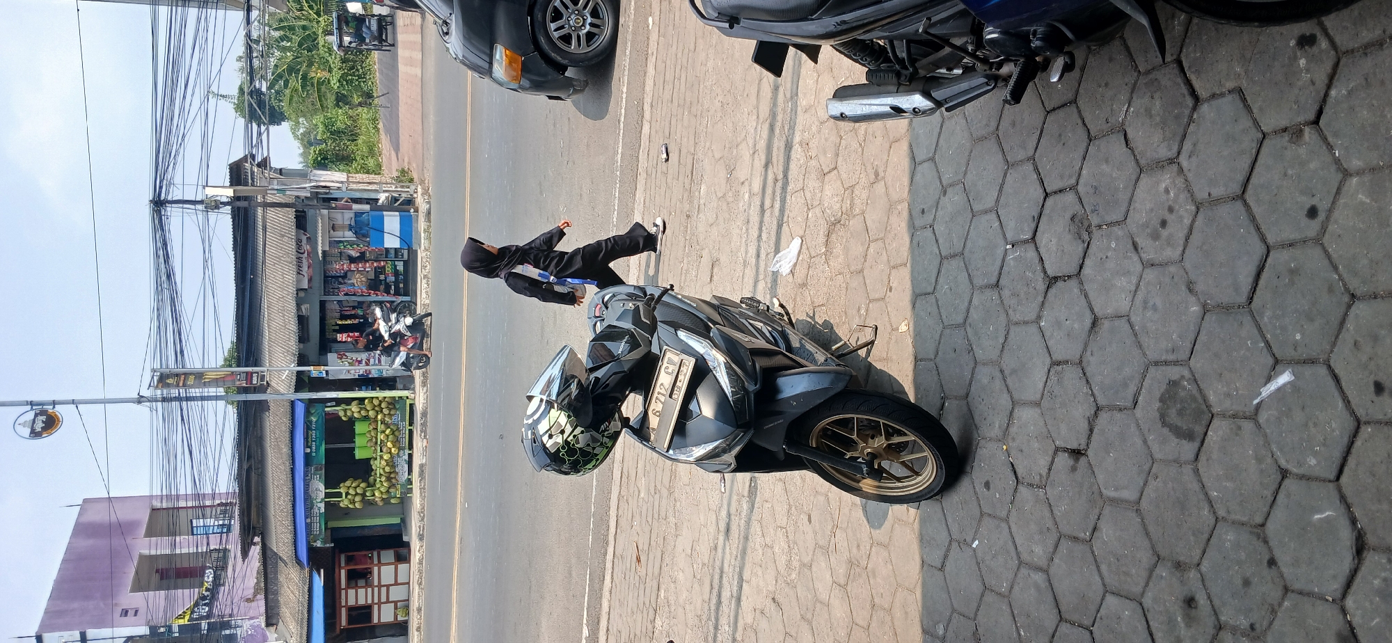In Baros, Indonesia, a child wearing a black hijab and a black outfit walks on a sidewalk paved with hexagonal concrete blocks. A black motorcycle with a silver and black helmet resting on the handlebars is parked in the foreground, facing slightly to the left. The motorcycle's license plate reads "A 6712 CV". In the background, a black van is visible on the road to the right. Across the street, there are several shops with awnings. One shop displays a large quantity of green coconuts. The sky is clear and blue, suggesting a sunny day. The scene depicts an everyday street view with traffic, pedestrians, and commercial establishments.