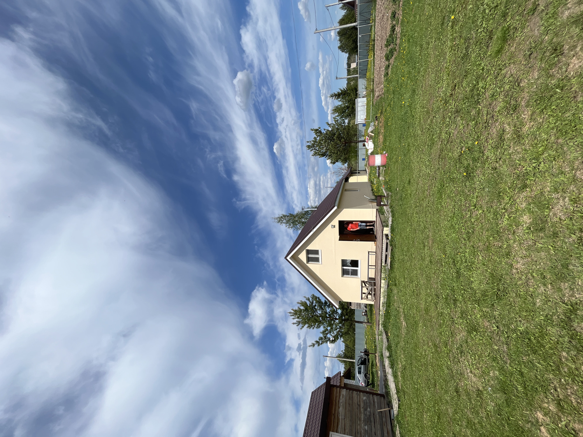 The scene depicts a rural residential setting in Гаревая, Russia, under a sky with streaky clouds. A single-story, beige house with a brown roof is prominently featured. A person wearing a red shirt is standing in the doorway of the house. A patch of green grass covers the foreground and extends to the right side of the image. A wooden structure with a brown roof is partially visible in the lower left corner. A silver car is parked behind a tree. A fence runs along the back of the property.