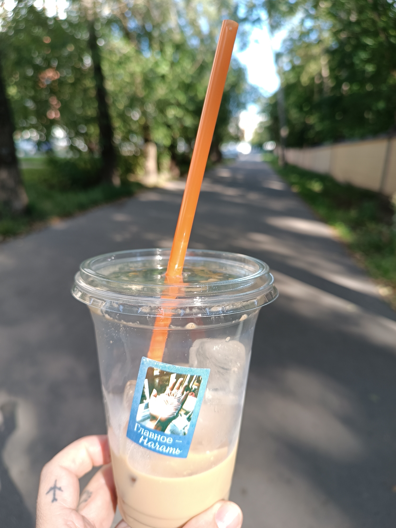 In Zhukovsky, Russia, on a paved path lined with trees and a yellow fence, a person holds a clear plastic cup filled with an iced beverage. An orange straw extends from the cup, which also has a blue sticker on its side with Russian text that reads "Главное — Память" (Mainly - Memory) and an image of an open hand with birds. A small airplane tattoo is visible on the person's thumb. The background is blurred, suggesting a shallow depth of field, with sunlight casting shadows on the path.
