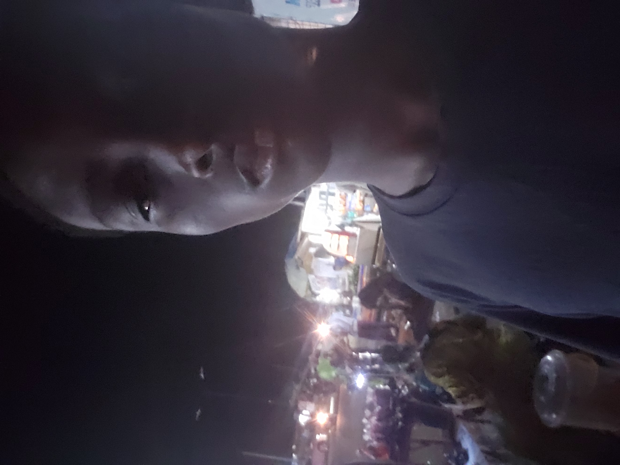The media file captures a dark, nighttime scene, likely a street or market in Ibadan, Nigeria, from a first-person perspective. The primary subject is a person in the foreground, with their face partially visible on the left side of the frame, looking slightly upwards and to the right. They are wearing a dark-colored t-shirt. Due to extremely low light, their features are largely obscured, but one eye is discernible, reflecting some of the background light.
The background is illuminated by numerous artificial light sources, creating strong glares and a busy atmosphere. These lights suggest the presence of stalls, shops, or street vendors. Indistinct figures of other people can be seen moving or congregating in the mid-ground, suggesting a lively environment. Several motorcycles are visible amidst the crowd, typical for an urban Nigerian street scene. In the bottom right corner, a clear plastic bottle is partially visible. The overall impression is of a bustling night market or street activity.
The time of day is clearly night, characterized by deep shadows and the reliance on artificial lighting, which casts bright flares across the frame. No specific weather conditions are discernible. There is no legible text visible in the image. The person in the foreground appears to be taking a selfie or a close-up shot, capturing the ambiance of the nighttime street behind them.