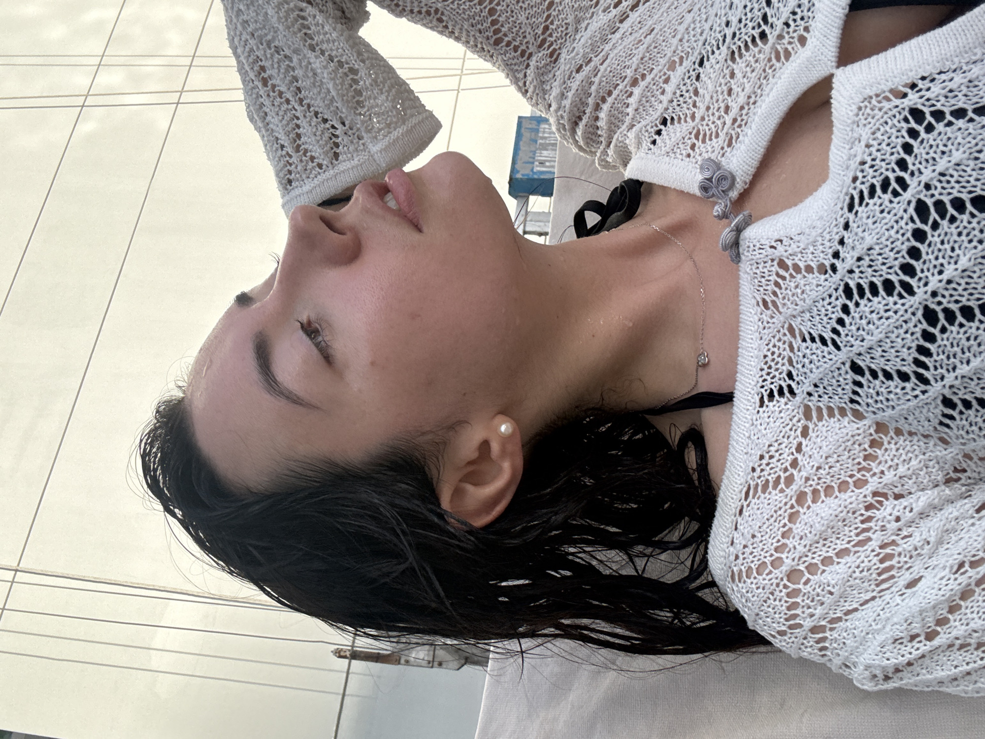 A woman is lying on a surface in Business Bay, United Arab Emirates. She has dark, wet hair and is wearing a pearl earring. She is wearing a black top and a white, open-knit cardigan with decorative clasps. A delicate necklace is visible around her neck. The background shows a building with glass panels and what appears to be a construction lift.