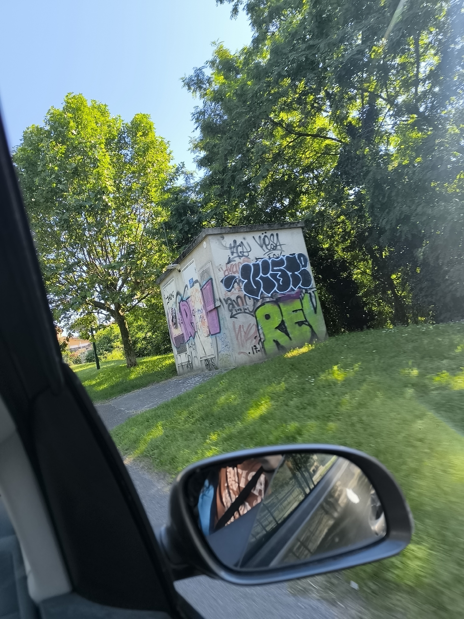The image depicts a view from inside a car, likely moving, in an outdoor setting possibly in Saint-Pierre-de-Bœuf, France (though this is unconfirmed from visual data alone). The main subject is a small, concrete utility building or structure heavily covered in graffiti. The graffiti includes words like "REVS" and other illegible tags in various colors. The building is positioned on a grassy slope, near a paved pathway. Background details include trees with dense green foliage, bathed in sunlight. The sky is visible and is a clear blue. A car's side mirror is prominently visible, showing a blurred reflection of the car's interior and the passing scenery. The interior elements suggest that a person is in the driver's seat (indicated in the reflection). The scene appears to be taken during the daytime under sunny weather conditions.