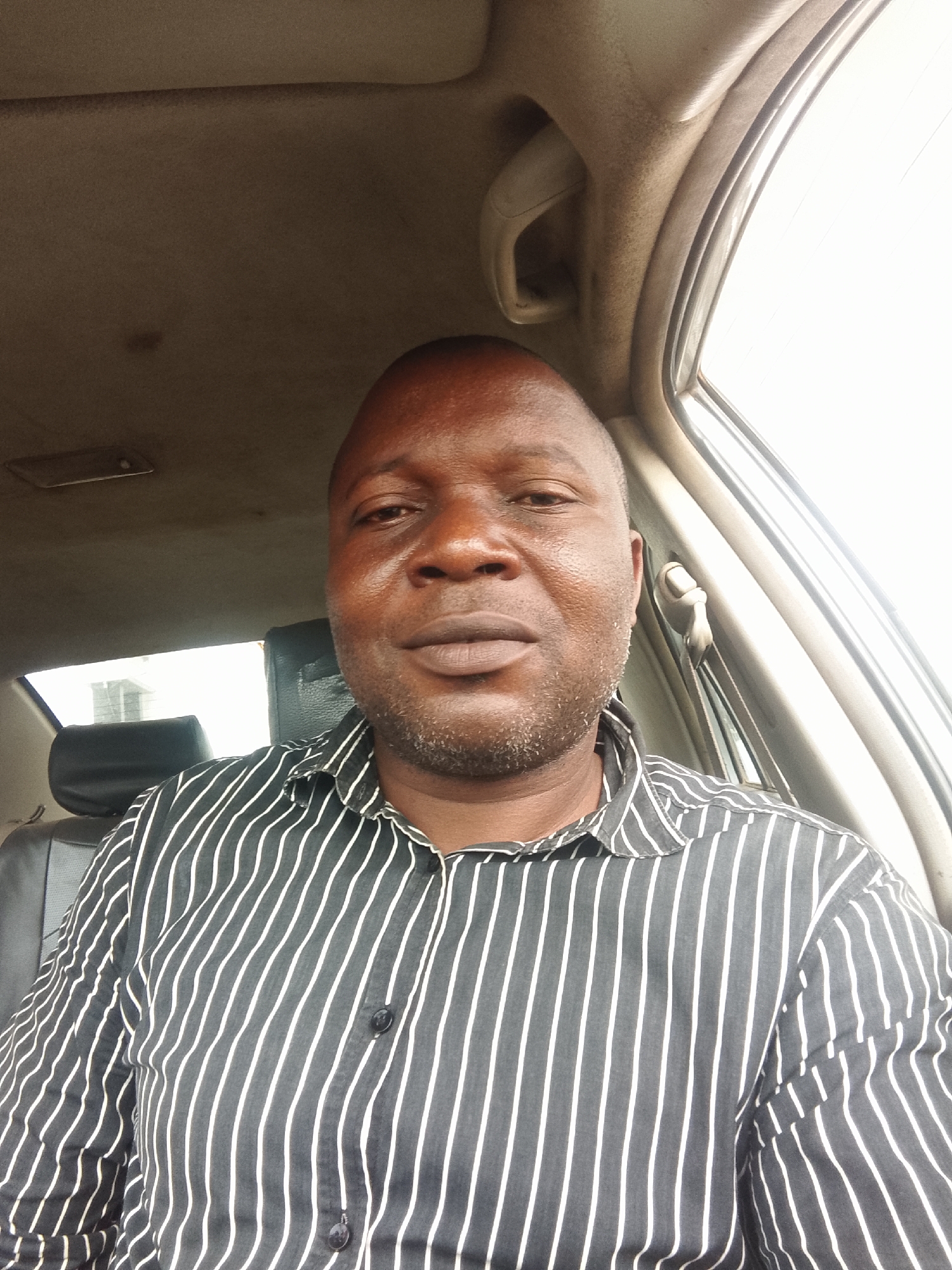 A medium-shot, high-angle selfie of a dark-skinned man in a black and white striped button-down shirt, sitting inside a car. The car's interior is visible, including the roof, window, and seat. Outside the window, there is a building. The location is likely Lokoja, Nigeria.