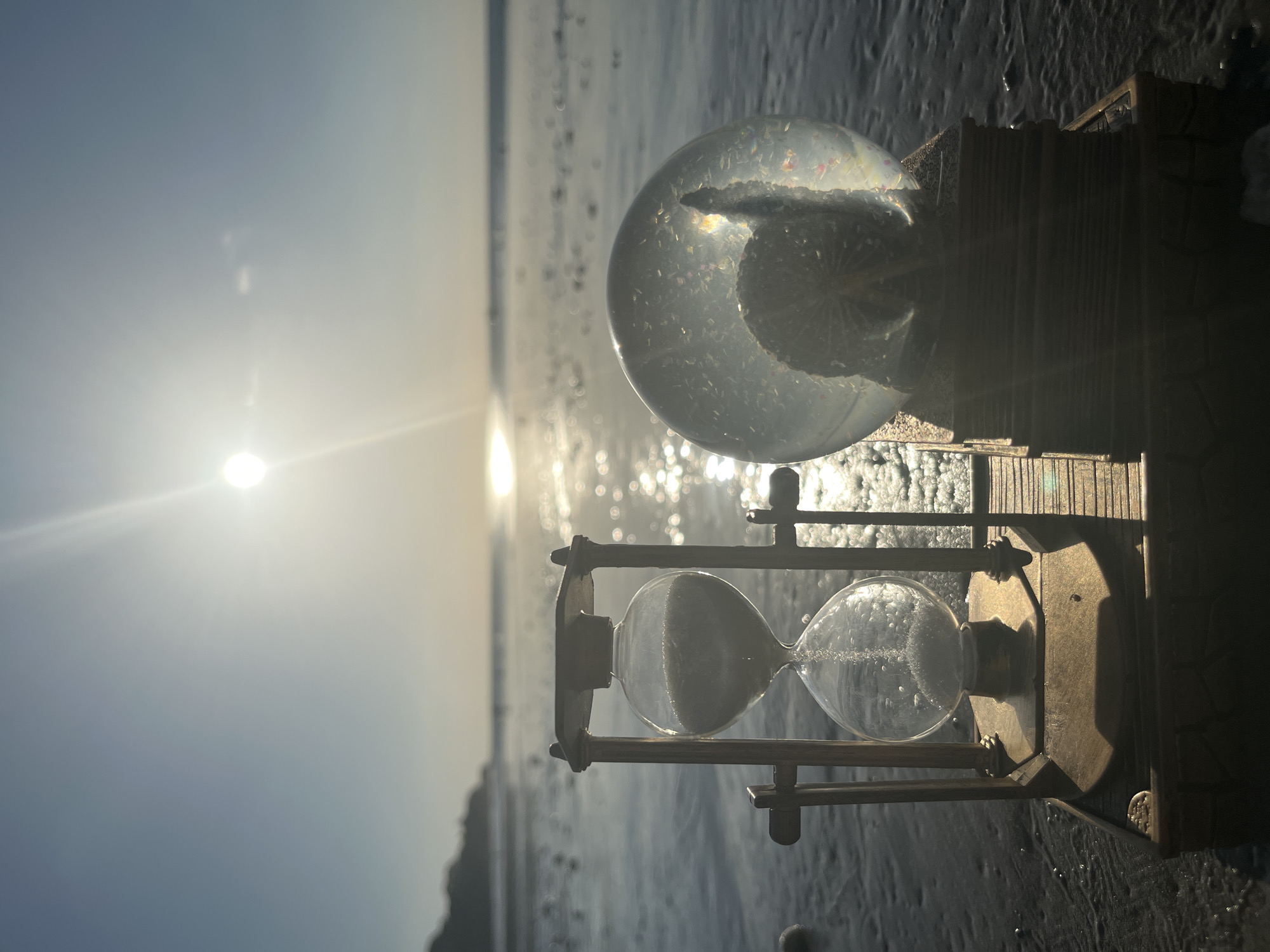 An hourglass and a snow globe sit on a wooden platform on a wet sandy beach. The hourglass is made of brass and glass, with sand in both bulbs. The snow globe is clear and contains a seashell and glitter. The sun is setting over the ocean in the background, casting a warm glow on the scene. The water is calm and reflects the light of the sun. The sand is dark and wet, with small waves lapping at the shore. There are no people visible in the image. The scene is serene and peaceful.