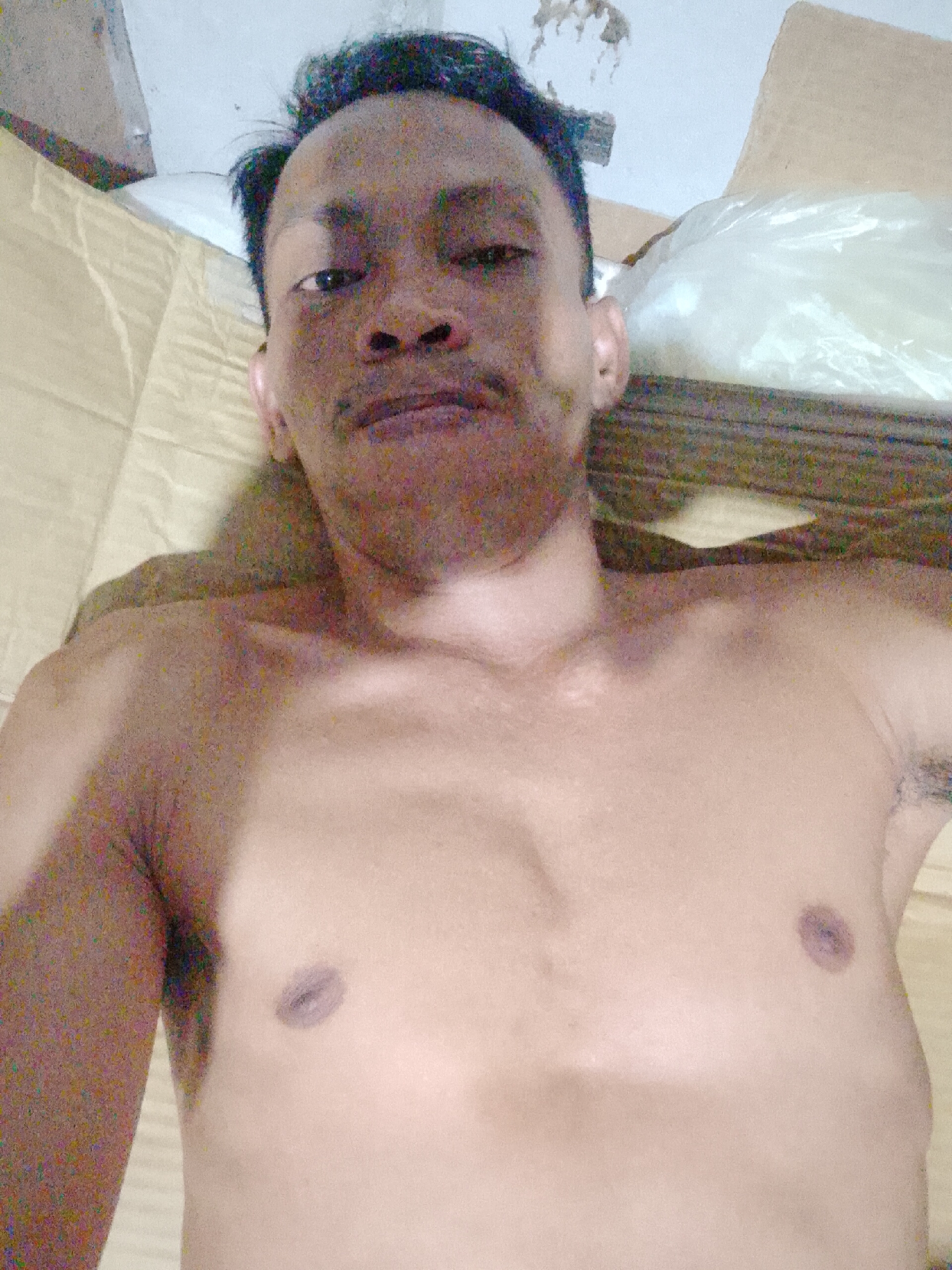 A shirtless adult male with dark hair, a mustache, and dark skin is lying on a brown cardboard surface. He is facing the camera. The location is Bukit Ketok, Indonesia. A plastic-covered object and more cardboard are visible behind him.