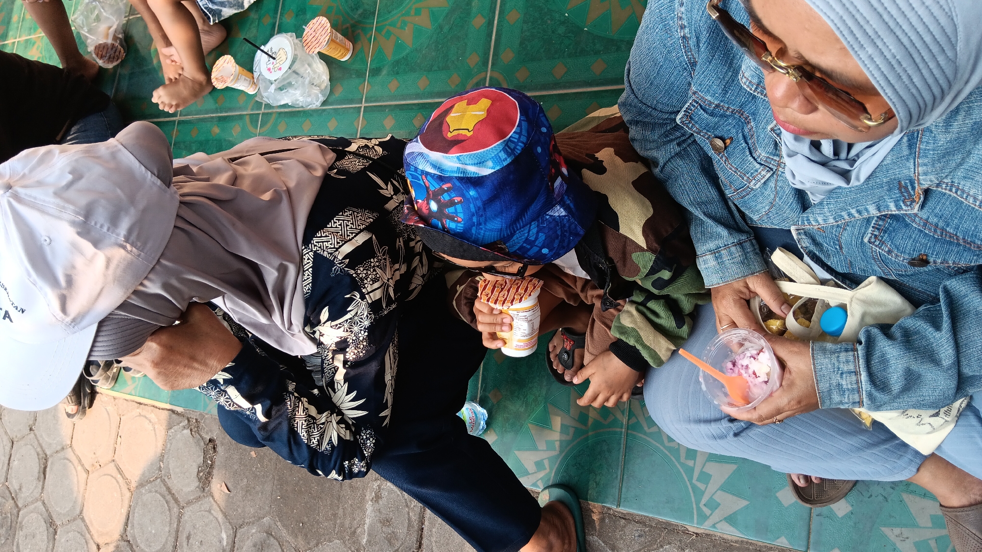 In Cerme, Indonesia, a group of individuals are seated outdoors on a patterned green tiled floor. The foreground is dominated by two people. A woman wearing a light blue denim jacket and light blue pants is seated, holding a clear plastic cup with an orange spoon and a light-colored dessert. Next to her, a child wearing a camouflage jacket and a blue hat with an Iron Man design is holding a cylindrical food container. To the left, another person is seated, wearing a dark blue patterned shirt and dark pants, with their head covered by a light gray hijab and a white baseball cap. In the background, other individuals, including children, are visible, and there are several disposable cups and plastic bags scattered on the ground. The environment suggests a casual outdoor gathering, possibly a park or a public rest area, with paved sections and greenery hinted at on the left side of the frame.