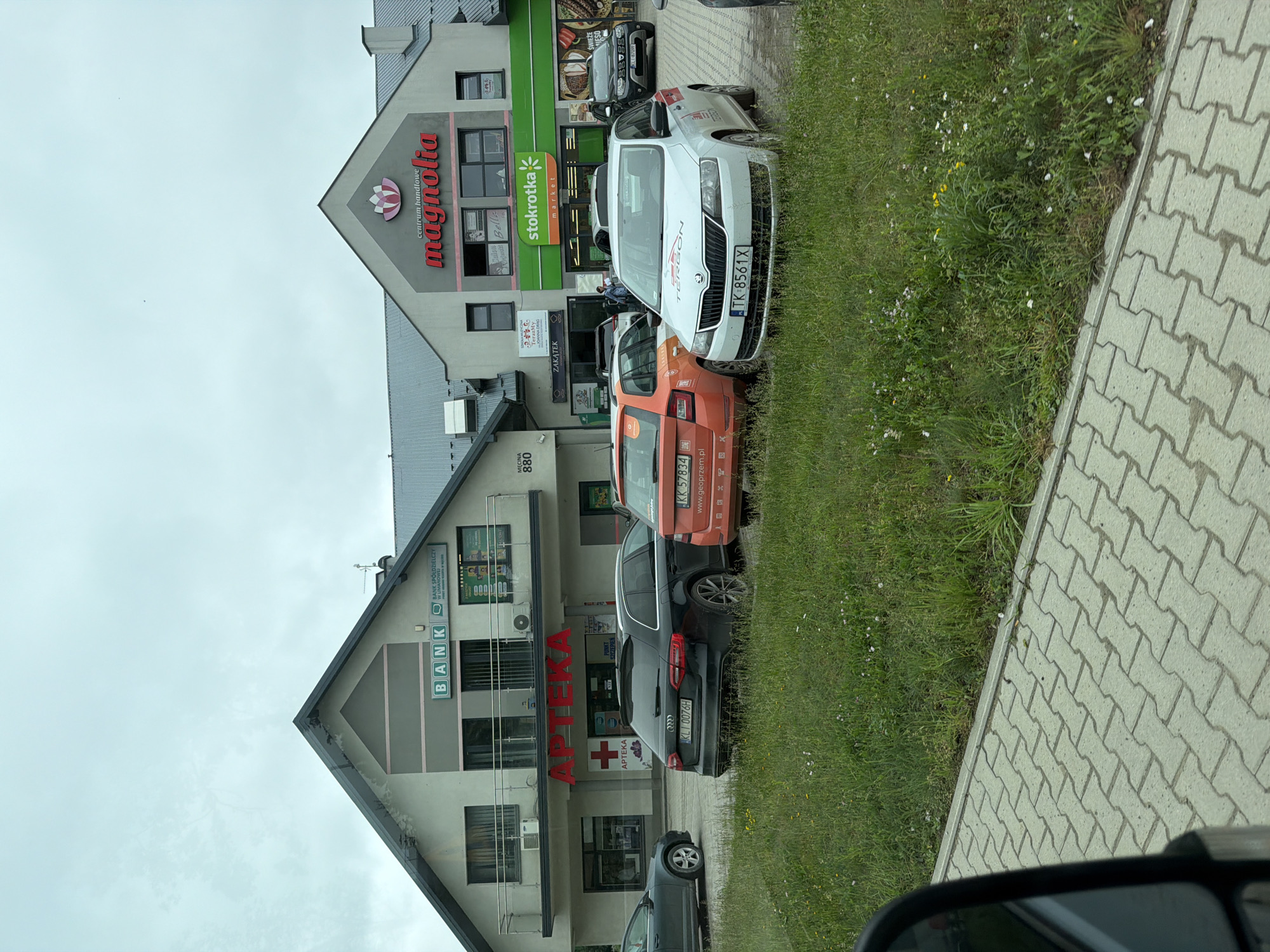 A medium shot captures a commercial building complex in Męcina, Poland, under an overcast sky. The building features prominent signage, including "BANK" and "APTEKA" on one section, and "centrum handlowe magnolia" and "stokrotka market" on another. Several cars are parked in front of the building and on the adjacent paved area. The foreground shows a section of a paved walkway and a strip of green grass with scattered wildflowers.