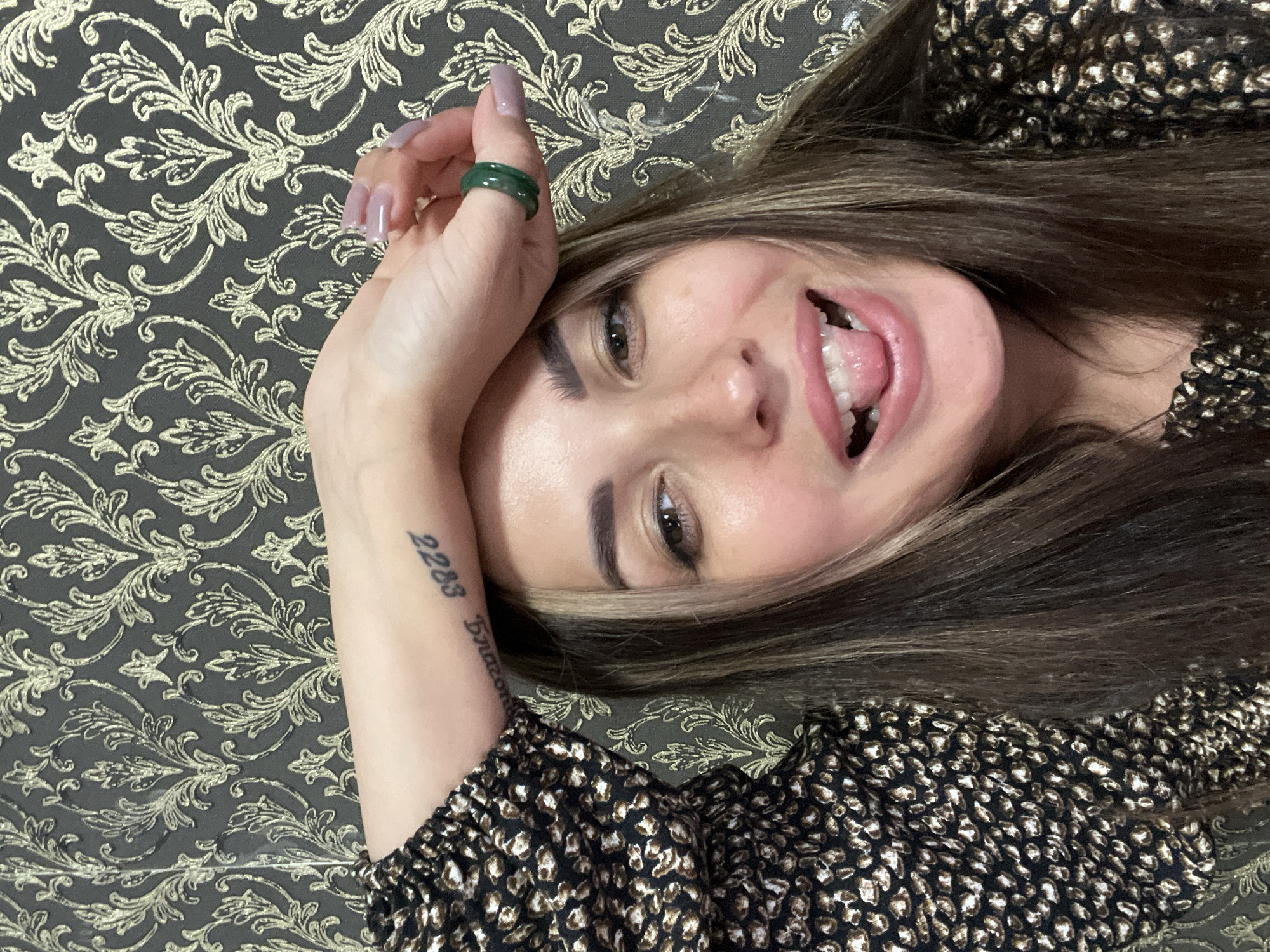 A medium close-up selfie shows a young woman in the foreground with her arm resting on her forehead. She has long brown hair with blonde highlights and is wearing a black and gold leopard-print top. She is sticking her tongue out and smiling playfully. A green ring is visible on her ring finger, and a tattoo with the numbers "2283" is visible on her forearm. The background is a dark gray wallpaper with a gold floral pattern. The image is taken in an indoor setting.