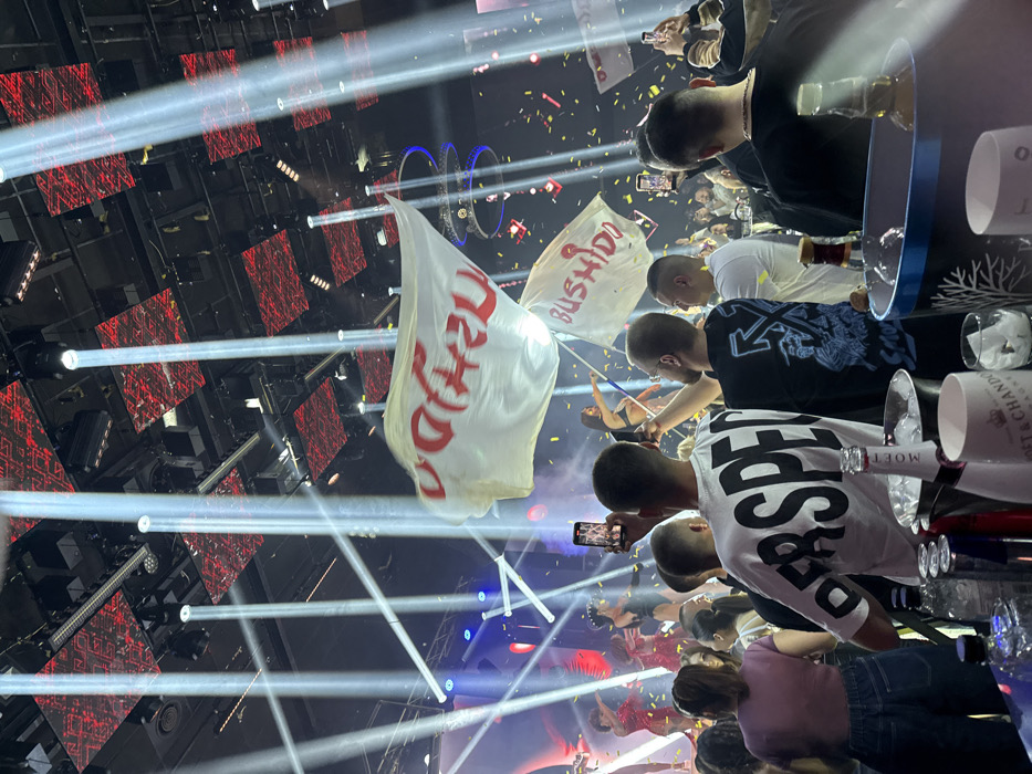 An indoor entertainment venue in Plovdiv, Bulgaria, is depicted during an active event. The scene features a crowded audience facing a brightly lit stage. Overhead, the ceiling displays red patterned panels and numerous spotlights emit strong white beams, creating visible light shafts through the air. Gold confetti is actively falling across the venue.
In the foreground, a bar surface or table holds various liquor bottles, including a chilled bottle of Moët & Chandon champagne in an ice bucket, alongside a blue ice bucket and several clear spirit bottles and glasses.
The midground is populated by numerous individuals, primarily observed from behind. Many attendees are holding smartphones aloft, actively recording or photographing the performance. A man in a white t-shirt with "DERSPE" visible on the back is central, holding up a phone. To his right, another man in a black t-shirt with a blue graphic also records with a phone.
On stage, multiple performers are visible under blue and red stage lighting. Several female performers, some in red sparkling outfits and others in black attire, are positioned on the stage. One performer in a black two-piece outfit is seen waving a white flag. Two large white flags with red stylized text, reading "BUSHIDO" (partially visible on the more prominent flag) are actively being waved, one by a performer and another by an attendee in the midground. The overall atmosphere suggests a high-energy performance or concert.