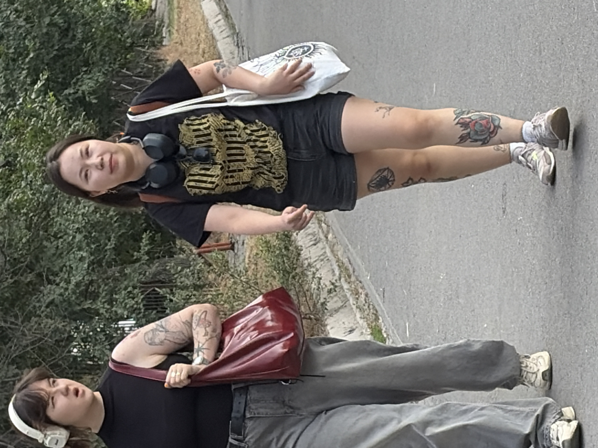 Two women are walking on a paved path lined with trees and shrubs in an urban environment. The woman on the left is wearing a black sleeveless top, grey baggy pants, and white headphones. She carries a shiny red purse on her left shoulder, and her arms have numerous tattoos. The woman on the right is wearing a black t-shirt with a gold graphic print, black denim shorts, and headphones around her neck. She has a white tote bag with a black graphic design slung over her right shoulder and also has visible tattoos on her arms and legs, including a rose on her left calf. Both women are wearing athletic shoes. The path appears to be a sidewalk or pedestrian walkway.