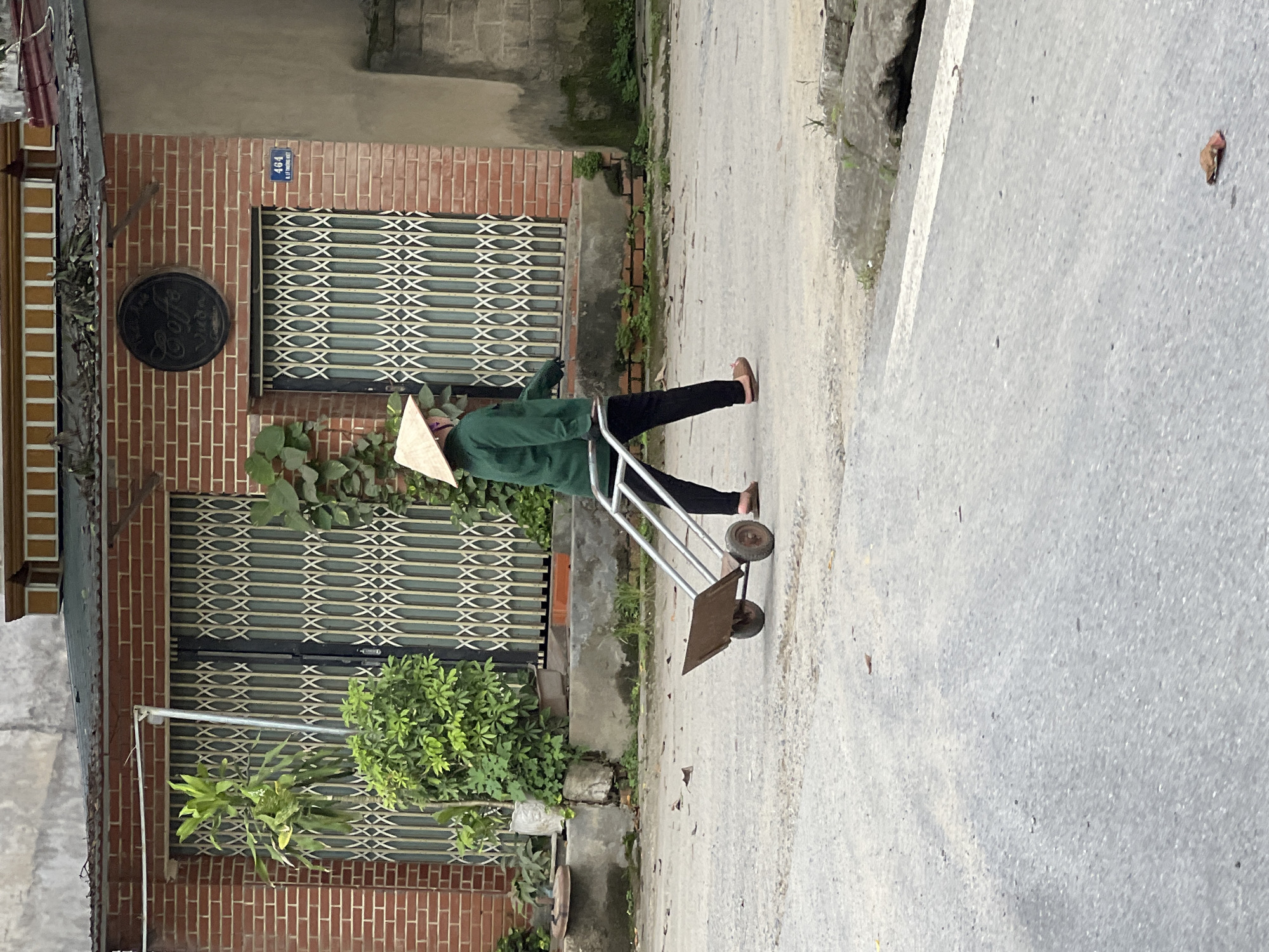 A person wearing a conical hat and a green jacket walks across a paved street in Hà Giang 2 Ward, Vietnam, pulling a two-wheeled handcart. The person is seen in profile, moving from left to right. The handcart is made of metal with a rusted platform and two spoked wheels. The background features a brick building with metal security gates and some greenery. A circular sign reading "Coffee" is affixed to the building wall. The street is partially wet and scattered with dry leaves.