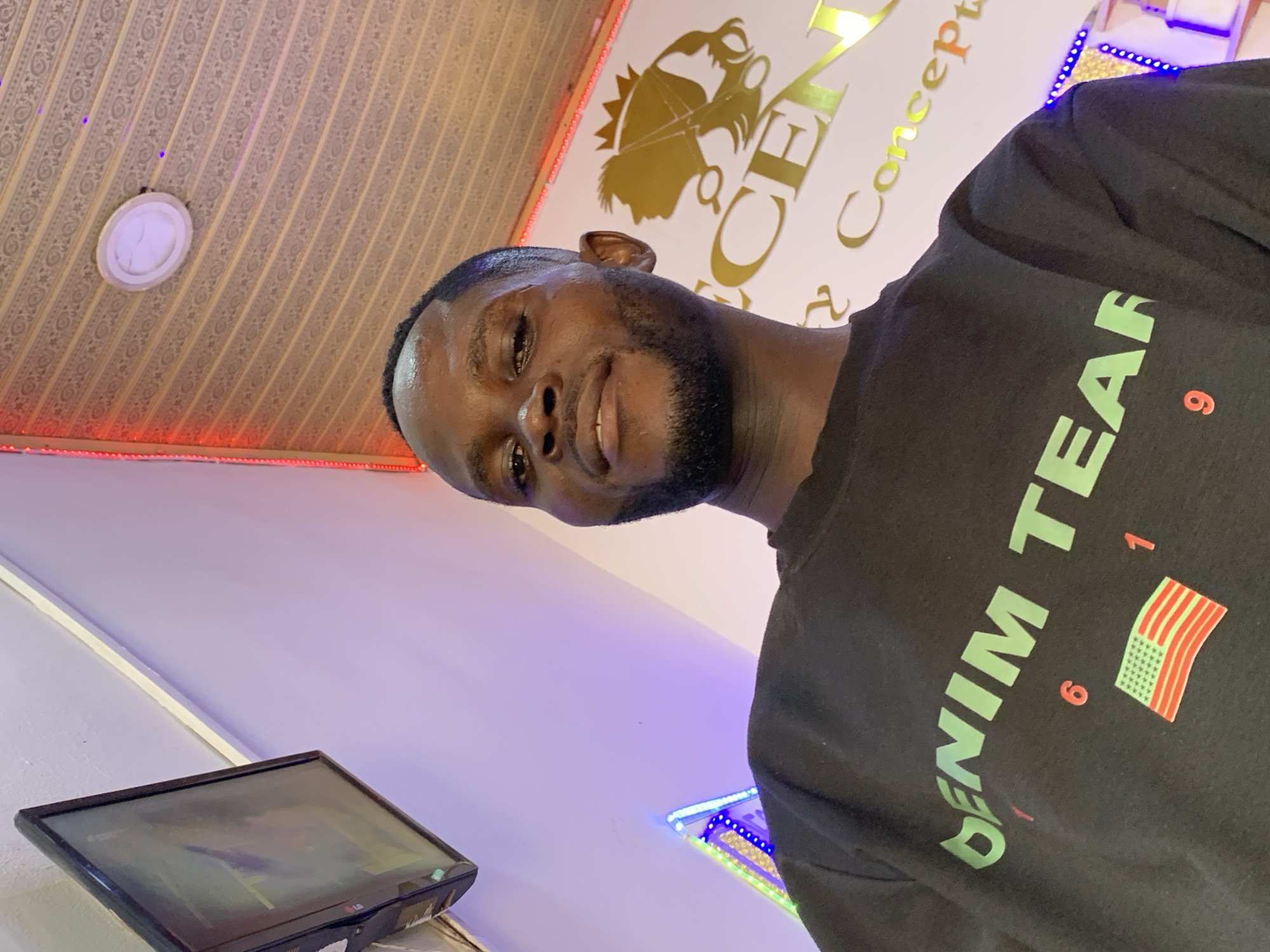 A medium-shot, low-angle photograph shows a man wearing a black t-shirt. The shirt has green lettering reading "Denim Team" with the number "619" and a stylized American flag to the right. The man has dark skin and a short, well-groomed beard. He is facing the camera with a slight smile.
The background includes a patterned ceiling with recessed lighting, a wall with a logo that reads "Concepts" in gold lettering, and a television set on a shelf. The wall is painted a light purple color. A string of lights are also visible. The location is Ikorodu, Nigeria.