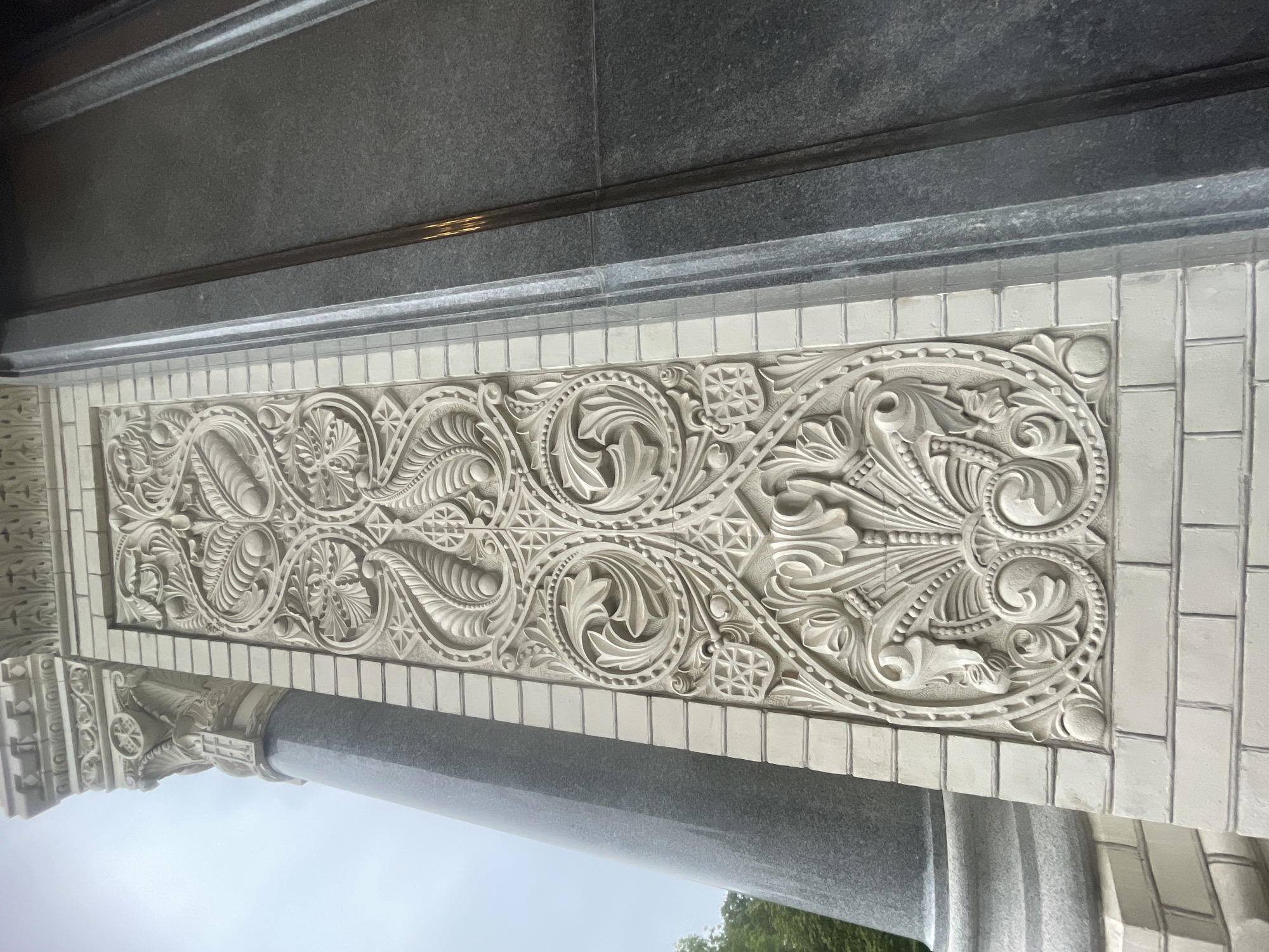 The image shows a close-up of the exterior architectural detail of the Naval Cathedral of Saint Nicholas in Kronstadt, Russia. The foreground features a decorative relief panel made of light-colored stone, possibly limestone or marble, set against a background of light-colored brickwork. The relief is highly ornate, depicting intricate floral motifs, scrolling leaves, and geometric patterns with a repetitive, symmetrical design. The panel is bordered by vertical bands of carved stone and narrow metal strips. To the left of the relief, a portion of a large, grey, fluted column is visible, suggesting it is part of a grand entrance or portico. To the right of the relief panel, a dark grey, polished stone facade is visible, likely granite, with visible seams indicating large stone slabs. The lighting suggests an overcast day, with soft shadows and muted colors. The perspective is upward-looking, emphasizing the scale and detail of the architecture. No people or actions are visible in the frame, focusing solely on the static architectural elements.