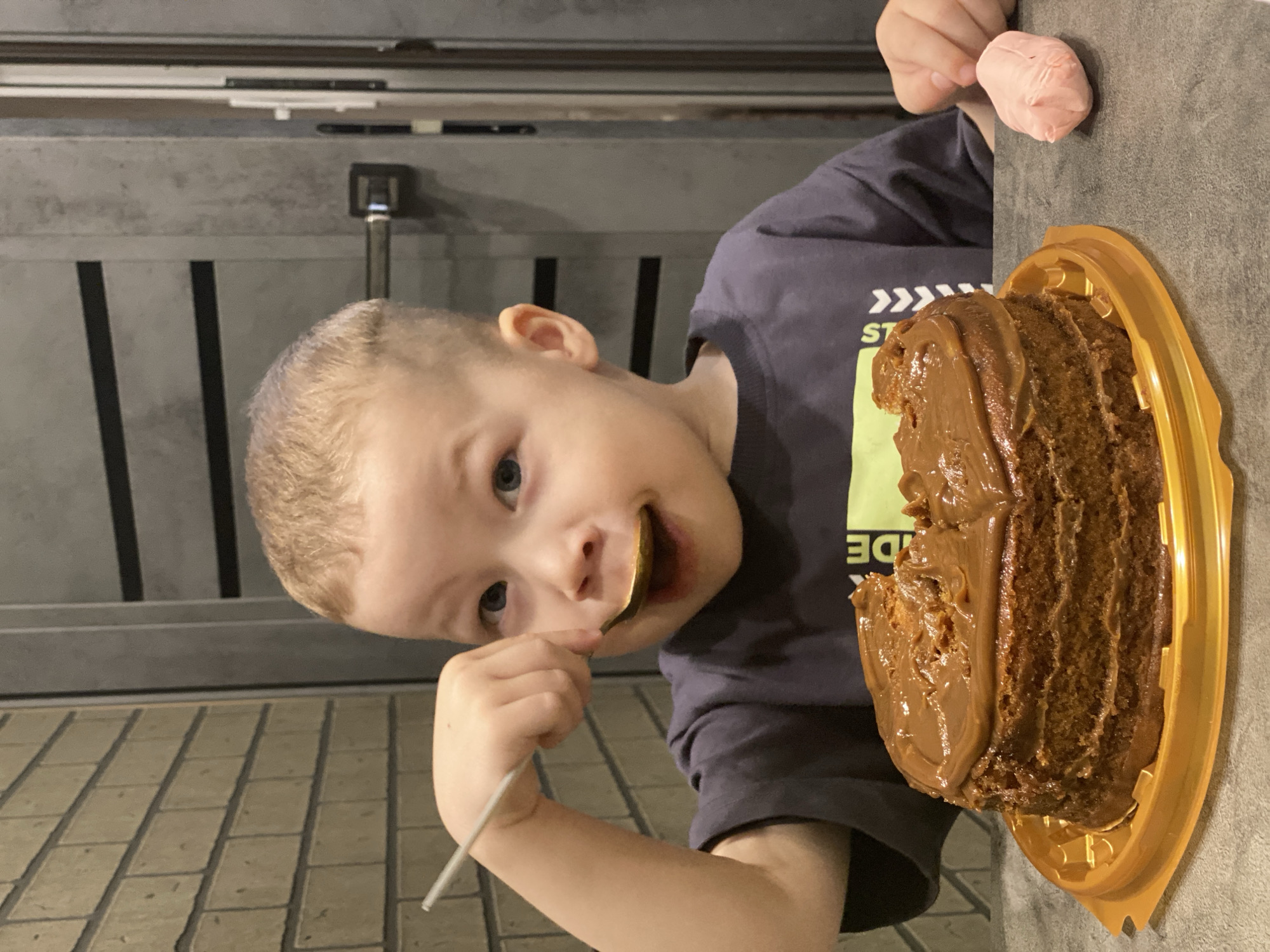 The image, which appears to be rotated 90 degrees counter-clockwise from its natural upright orientation, captures a delightful indoor moment in Sochi, Russia. A young, blonde-haired child with bright blue eyes is the central subject, positioned to the left of the frame (when upright). He gazes directly at the camera with a playful or contented expression, his mouth slightly smeared with frosting. He holds a golden spoon, still laden with some of the light brown frosting, near his mouth.
To the child's right, on a dark gray tabletop, sits a large, round chocolate cake. The cake is generously covered with a thick layer of light brown frosting, possibly dulce de leche, and is placed in a golden plastic container. A significant portion of the cake has already been eaten, indicating an ongoing moment of indulgence. In the upper right corner of the frame, a small, pink, irregularly shaped candy or marshmallow is held by an unseen hand.
The child is wearing a dark gray t-shirt, on which partial white and light green text is visible, reading "ST" and "DE". The background behind the child features dark gray walls or furniture with prominent vertical lines (which would be horizontal if the image were upright), and a lighter-colored floor with a repeating brick-like or tiled pattern. The scene conveys a joyful and casual event, likely a treat or celebration, under artificial indoor lighting.