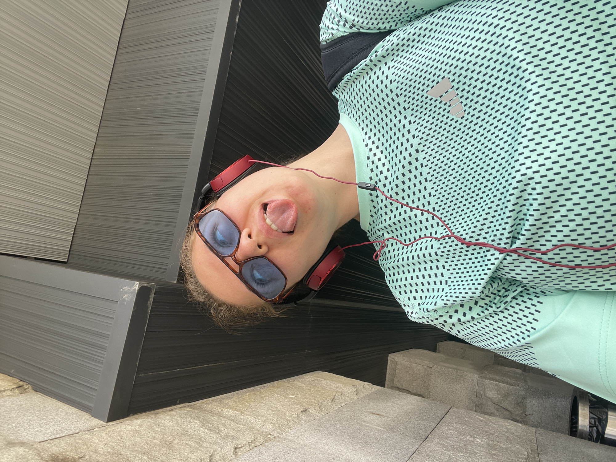 A person is seen from a low angle, appearing to be upside down. They are wearing sunglasses, headphones, and a patterned green shirt with a black sports bra underneath. Their tongue is sticking out. The background shows a gray and black panelled wall and gray steps. This location is in Moscow, Russia.