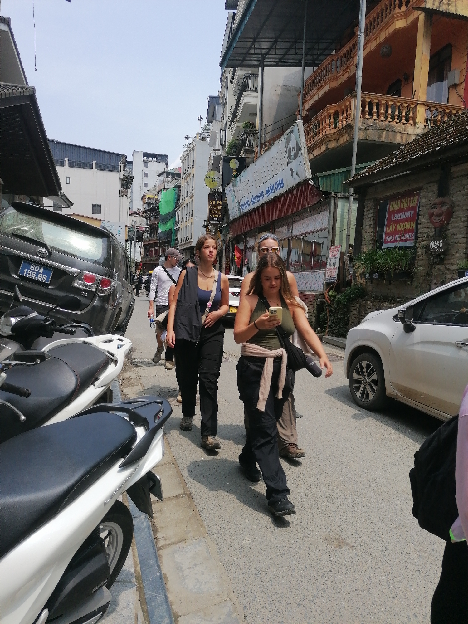 The image shows a street scene in Sa Pa Ward, Vietnam. Three women are walking down the street. The first woman is wearing a blue tank top with a black vest and black pants, and the second woman is wearing a green tank top with a beige jacket tied around her waist and black pants. The third woman is wearing a white headband and tan pants. The first woman is looking ahead, while the second woman is looking at her phone.
In the background, there are buildings and parked cars and motorbikes. Street signs include, "SA PA CLOVER HOTEL" and "LAUNDRY."