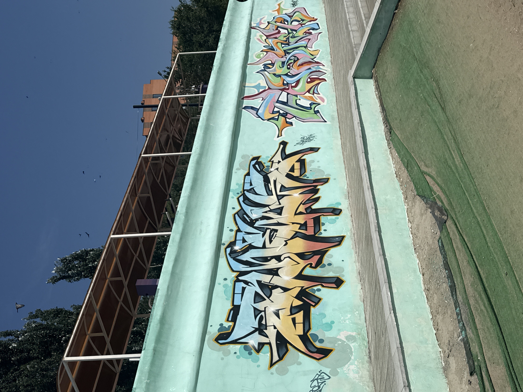 The image depicts a section of a green-painted concrete wall adorned with colorful graffiti. The graffiti on the left is larger, featuring stylized block letters with a gradient of light blue, yellow, and pink, outlined in black and accented with white highlights. To its right, smaller, more abstract graffiti in a vibrant palette of green, pink, orange, and blue is present. The wall is situated outdoors, likely in a public space, given the presence of a paved walkway and steps bordering it. Above the wall, a covered structure with a brown roof supported by white pillars is visible, suggesting it might be a seating area or shelter. Trees line the background, and the sky above is clear blue with several birds in flight. The ground below the wall appears to be a mix of dirt and green netting or turf. The overall scene suggests an urban environment with elements of public art and outdoor amenities. This location is identified as being in Pavlodar, Kazakhstan.