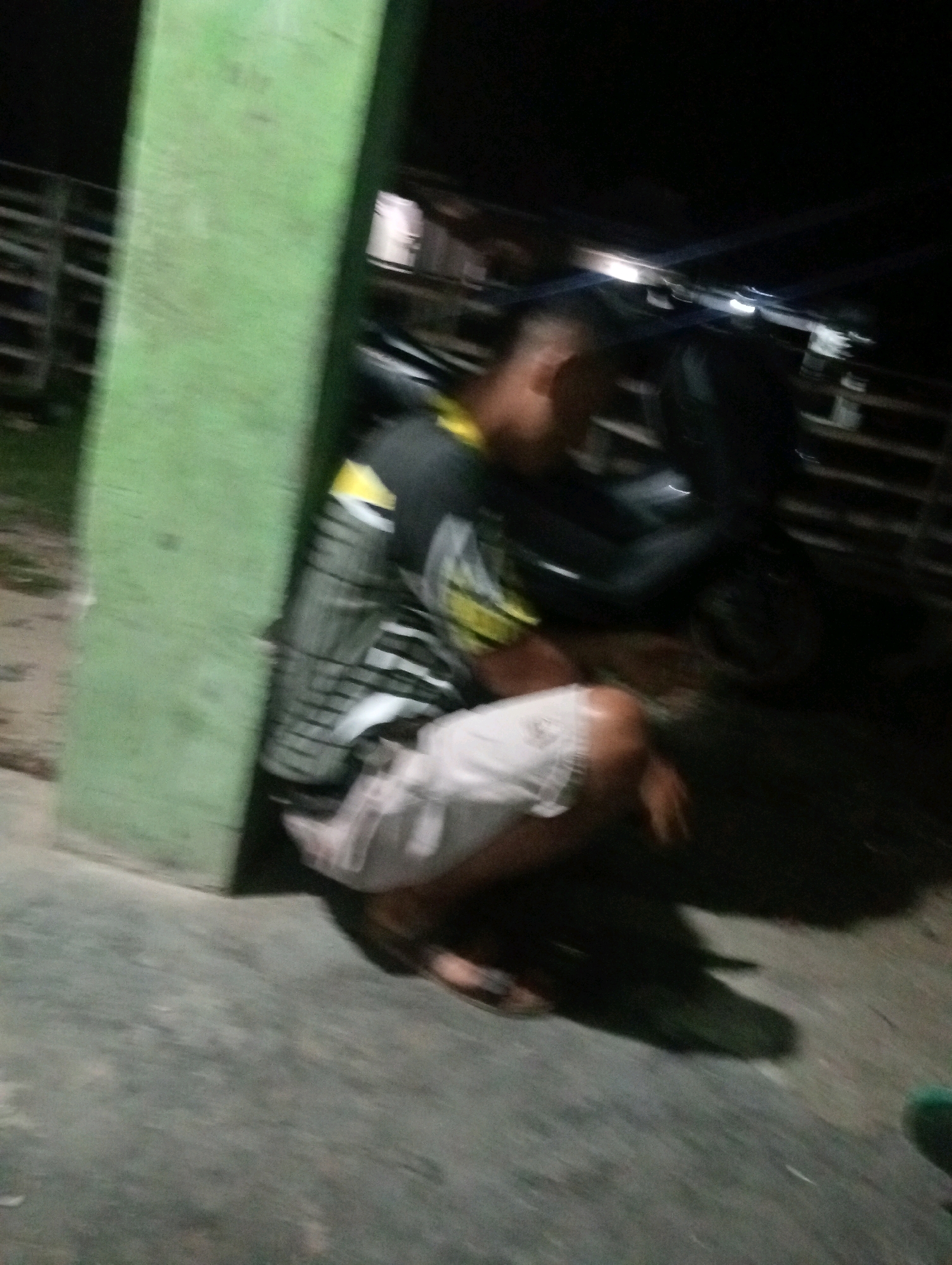 A person is crouched on the ground next to a green pillar in an outdoor, dimly lit environment, possibly at night. The person is wearing a dark shirt with a yellow and white pattern and light-colored shorts. A dark motorcycle is partially visible behind the person. The ground appears to be concrete or asphalt. In the background, there are structures that resemble fences or railings. The image is blurry, making it difficult to discern finer details. The scene is consistent with an urban or semi-urban setting.