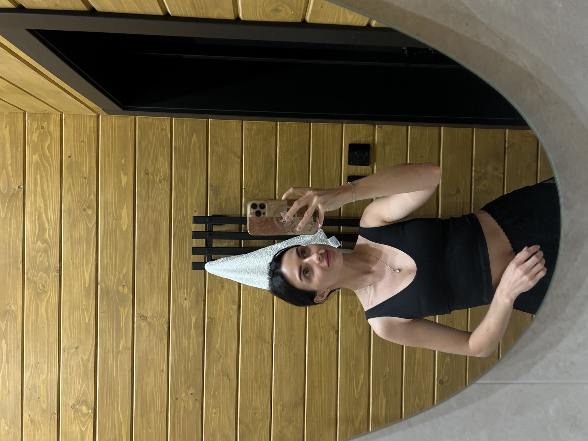 A young woman with dark hair, wearing a black tank top and black shorts, takes a mirror selfie in a bathroom. The bathroom walls are paneled with horizontal wooden planks. A white towel hangs on a black rack behind her. To her right, a black door frame is visible. On the wall to her right, there is a small digital display showing "25". The floor appears to be tiled. The woman is holding a smartphone with a pink case decorated with a Hello Kitty sticker in her right hand, and her left arm is resting at her side. She is wearing a silver necklace with a clear pendant and a thin silver bracelet on her left wrist. The overall setting suggests a modern, possibly minimalist, bathroom in a home or hotel. There is no specific information or visual cues in the image to indicate the location in the city of Stradch, Ukraine.