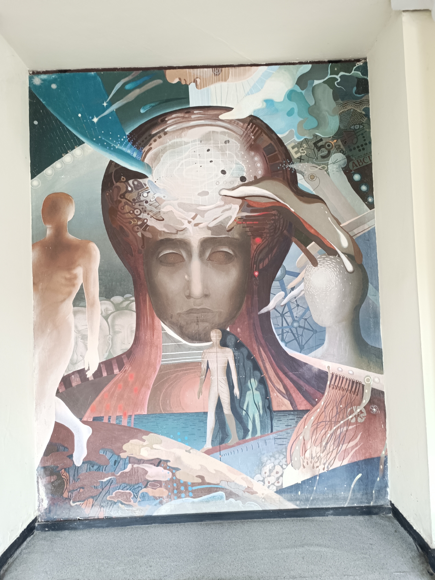 The image shows a mural on a wall, possibly indoors, in Sofia, Bulgaria. The mural depicts a complex, surreal scene featuring human figures and abstract elements.
A large, central face dominates the composition. The face is androgynous, with closed eyes and a solemn expression. Above the face, the head is represented as a complex structure resembling a circuit board or a neural network.
To the left of the face, a nude, pale figure stands with its back to the viewer. Behind it, faint, indistinct figures appear in the background.
On the right side of the mural, there is another stylized human head. A hand appears to be interacting with this head, possibly representing knowledge or communication.
The lower portion of the mural depicts a landscape with water and landforms. A smaller, nude figure stands on this landscape.
The mural incorporates elements of space, science, and the human body. Symbols, letters, and abstract patterns are also present. The style is reminiscent of surrealism and possibly influenced by themes of technology, consciousness, and the human condition.
The wall appears to be part of a building interior. The floor is gray and featureless, and the surrounding walls are painted white.