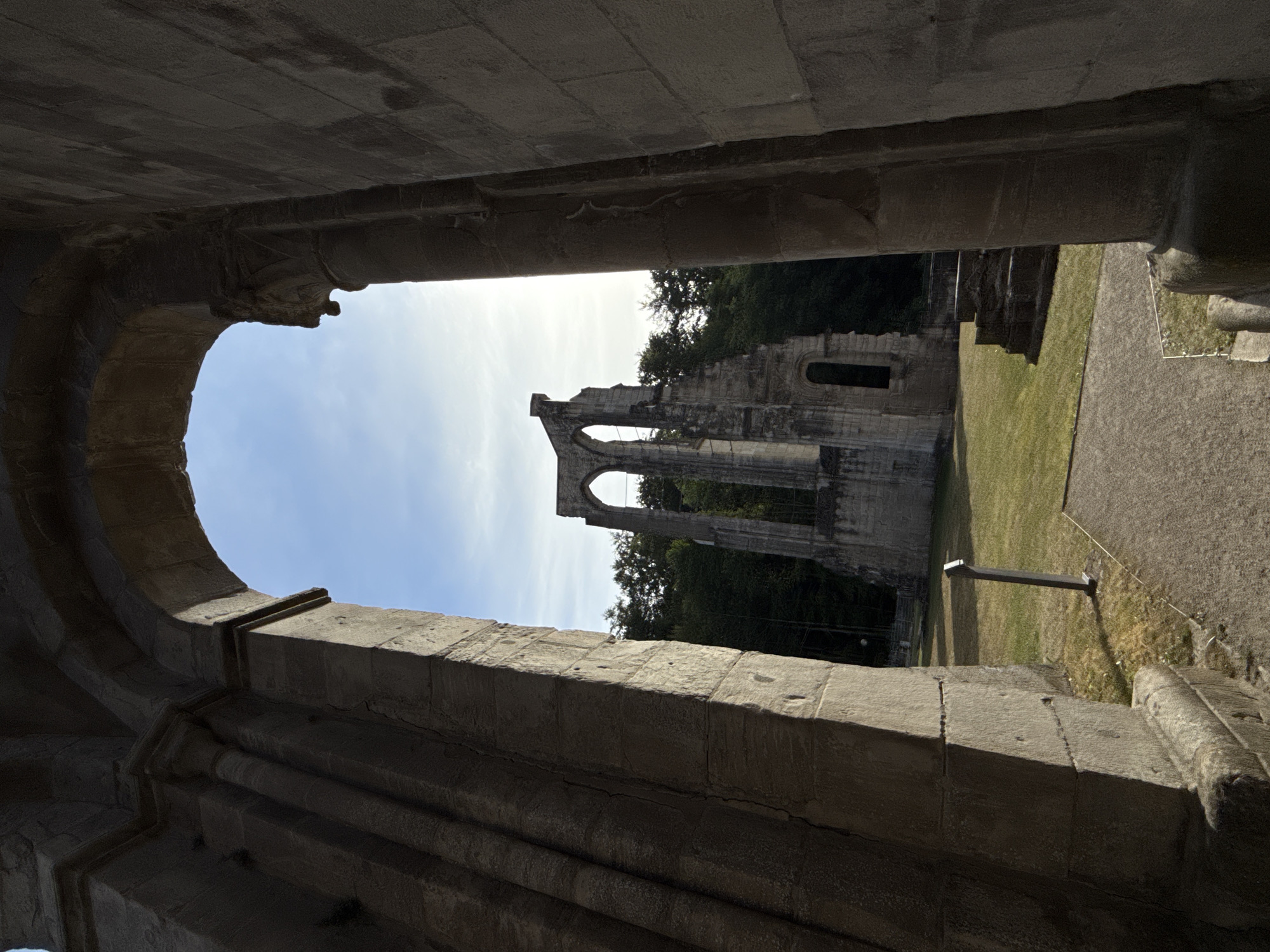 The image shows a view through a stone archway of the ruins of Walkenried Abbey in Walkenried, Germany. The ruins are made of large, weathered stone blocks. The foreground is dominated by the dark, shadowed interior of the archway, with detailed stonework visible on the left and right. Through the arch, the skeletal remains of the abbey's architecture are visible against a clear blue sky with some wispy clouds. These ruins consist of tall, crumbling stone walls with arched openings. The ground in front of the ruins is a grassy area with a gravel pathway leading towards it. There are no people present in the scene. The overall impression is one of historical grandeur and decay.