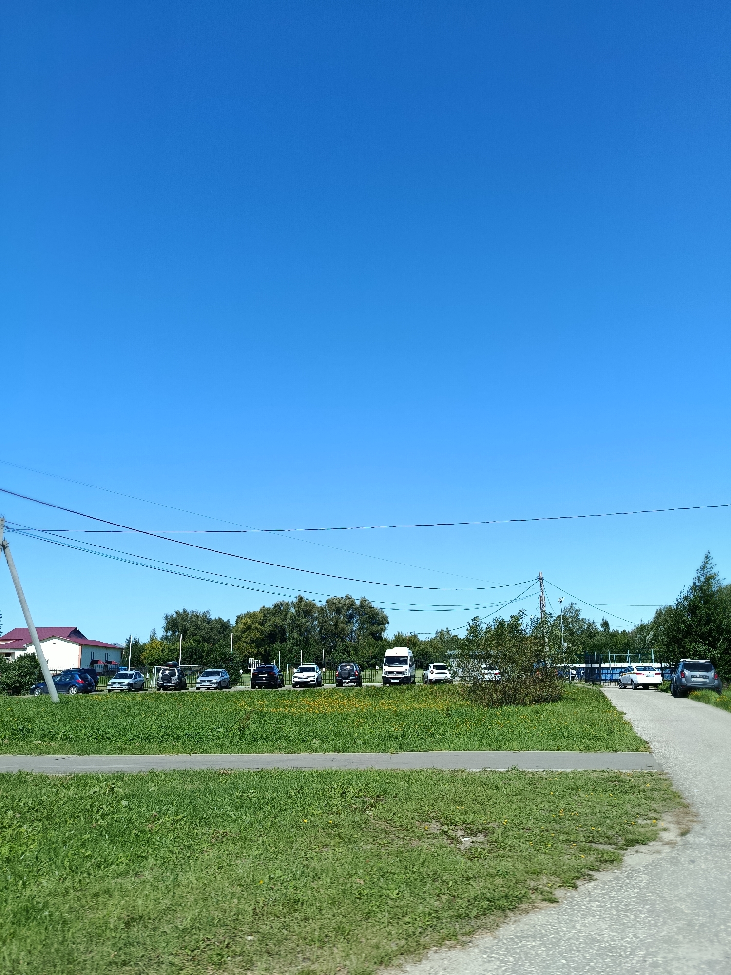 The image shows a street scene in Vereya, Russia. A paved road curves to the right in the foreground, bordered by a grassy area. Beyond the grass, a row of parked cars is visible, including a white van, several SUVs, and sedans. In the background, there are trees and buildings, including a red-roofed structure. Power lines are strung across the sky. The sky is clear and blue, indicating fair weather. There are no people visible in the image.
