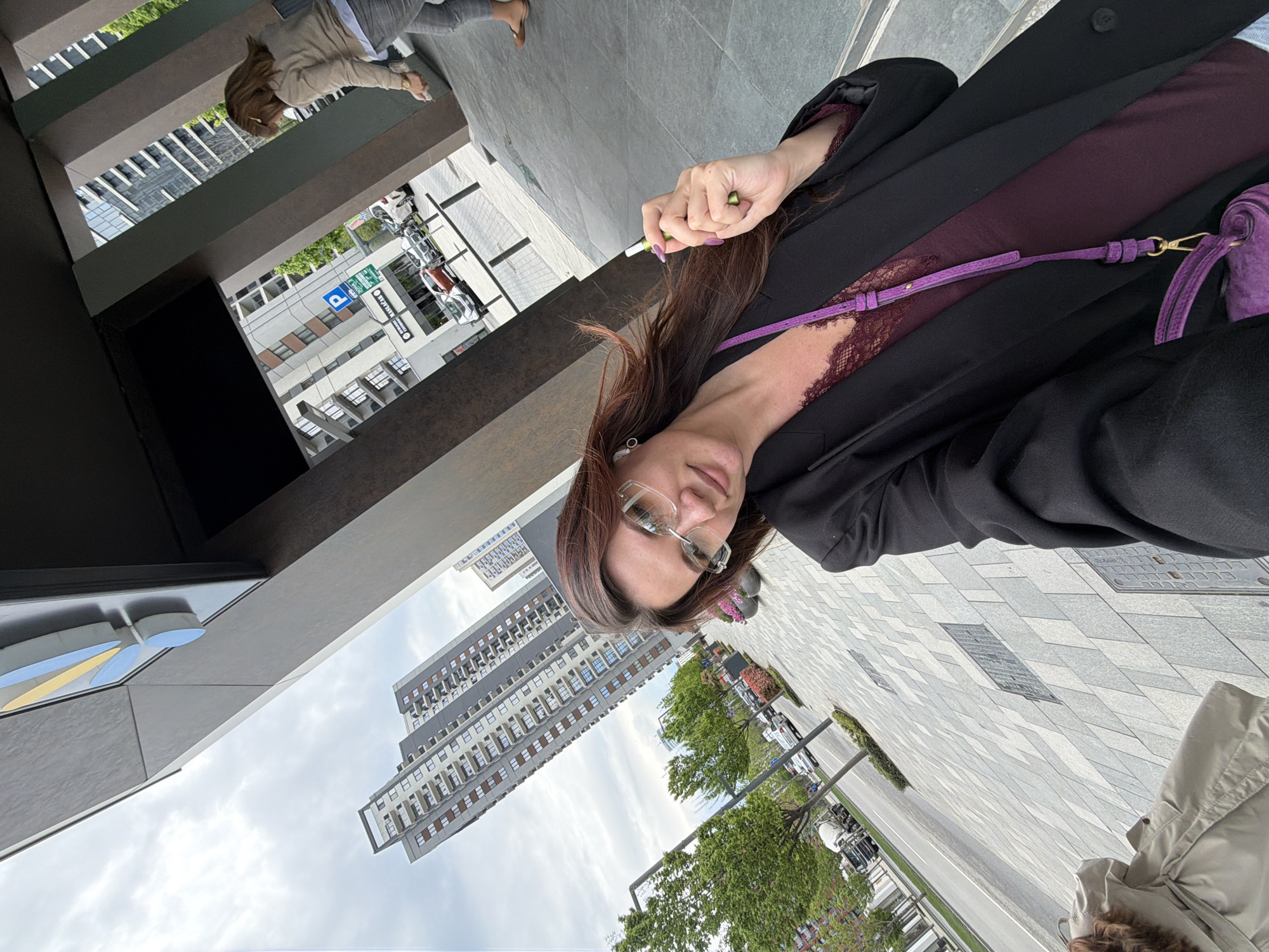 An urban selfie features a woman posing outdoors, captured from a slightly low angle. The image is rotated 90 degrees counter-clockwise.
The primary subject is a woman with long, dark brown hair, parted on the side, wearing clear-framed glasses. She looks towards the camera with a neutral expression. She is dressed in a black blazer or jacket over a dark purple top, which appears to have lace detailing around the neckline or shoulder. A thin purple strap, possibly from a handbag, crosses her chest. Her right hand is raised near her hair, holding a green and white pen-like object, and her fingernails are painted light purple.
The setting is a modern city environment. In the background, there are multi-story buildings with numerous windows, suggesting contemporary architecture. A paved area with a distinctive geometric tile pattern stretches out, leading to a street with parked cars and lush green trees. The sky is overcast. Another person in light-colored clothing is partially visible further back, appearing to walk.
Notable details include a blue and white parking sign in the middle ground, which clearly displays the letter "P" for parking and the word "BESPLATNO," meaning "FREE" in Serbian, confirming the location context of Belgrade, Serbia. Another partially visible sign reads "MAXX." The scene depicts a daytime urban ambiance, possibly on a cloudy day.