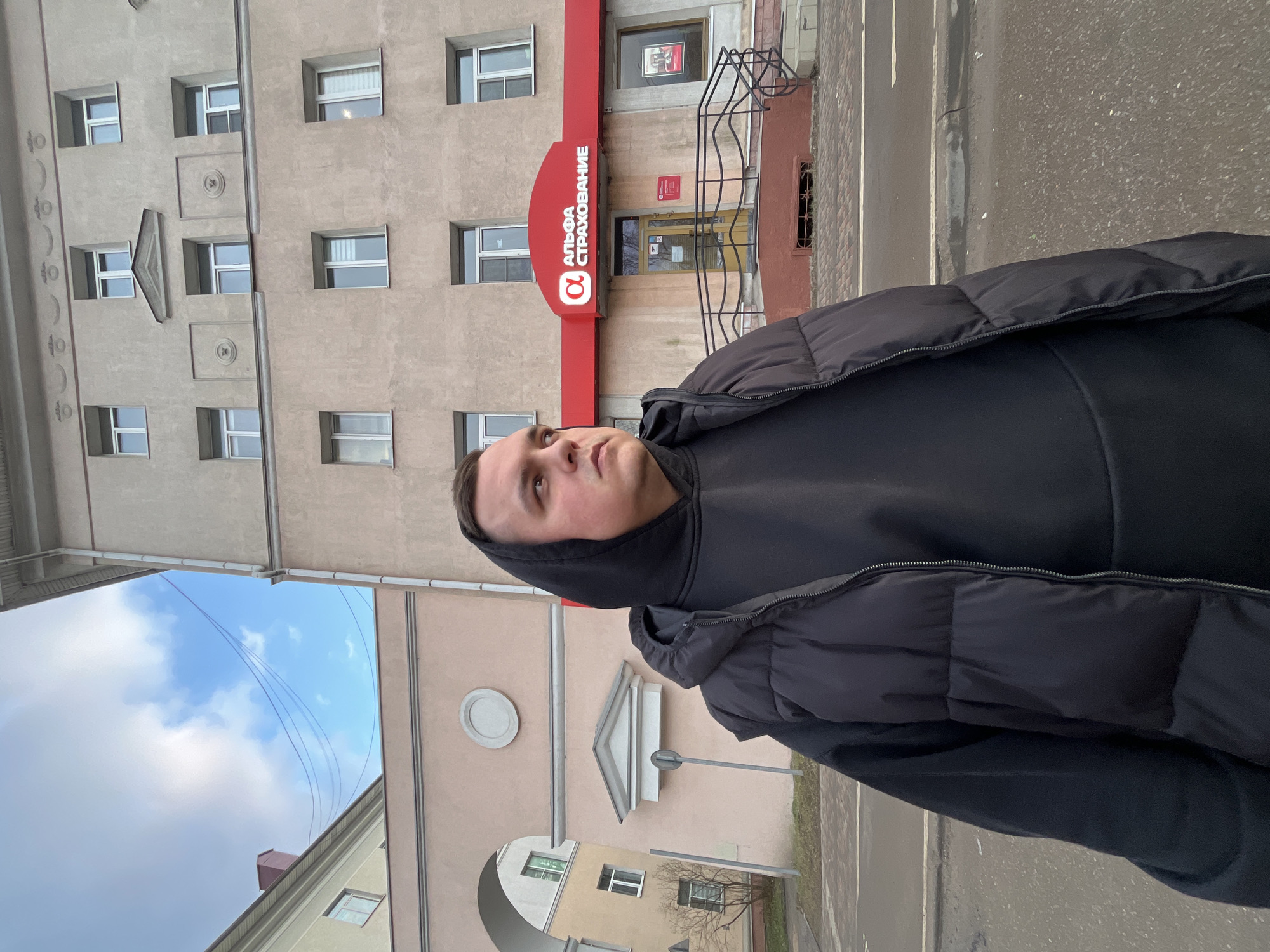 A man wearing a black hooded sweatshirt and puffy jacket stands on a street in Klin, Russia. Behind him is a building with the Alfa Strakhovanie insurance company logo. The weather appears overcast, and a sidewalk and road are visible. Buildings and a partially visible sky are in the background.