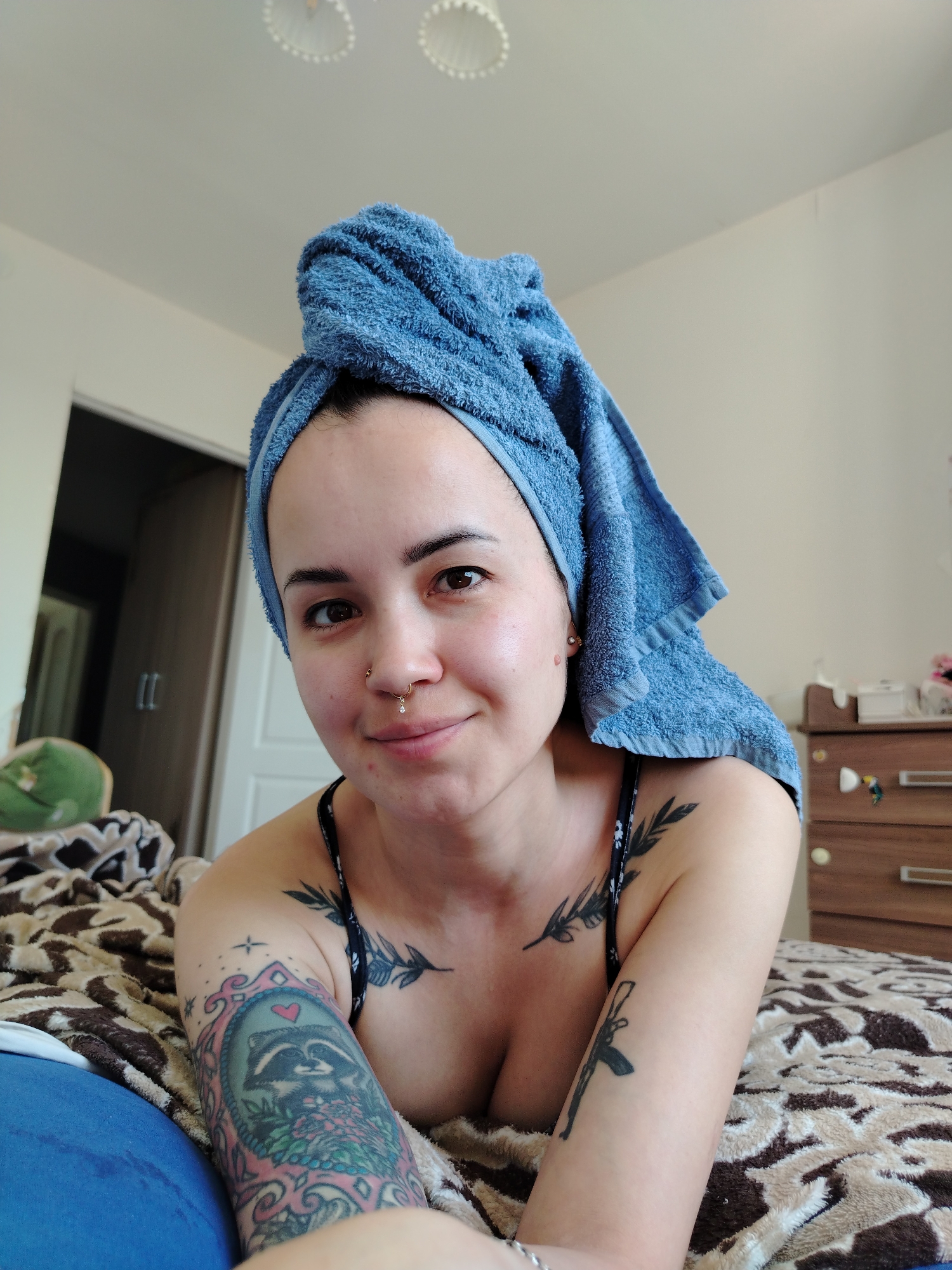 A woman is lying on a bed with a brown patterned blanket. She has a blue towel wrapped around her head. The woman has multiple tattoos on her arms and chest. Her right arm has a tattoo of a raccoon in a frame with flowers. The left arm has a tattoo of a gun. Her chest has two small tattoos of leaves. There is a door behind her, a dresser to her right, and a green stuffed animal on the left side of the bed. The photo does not depict the location in the city of Noviy, Russia.