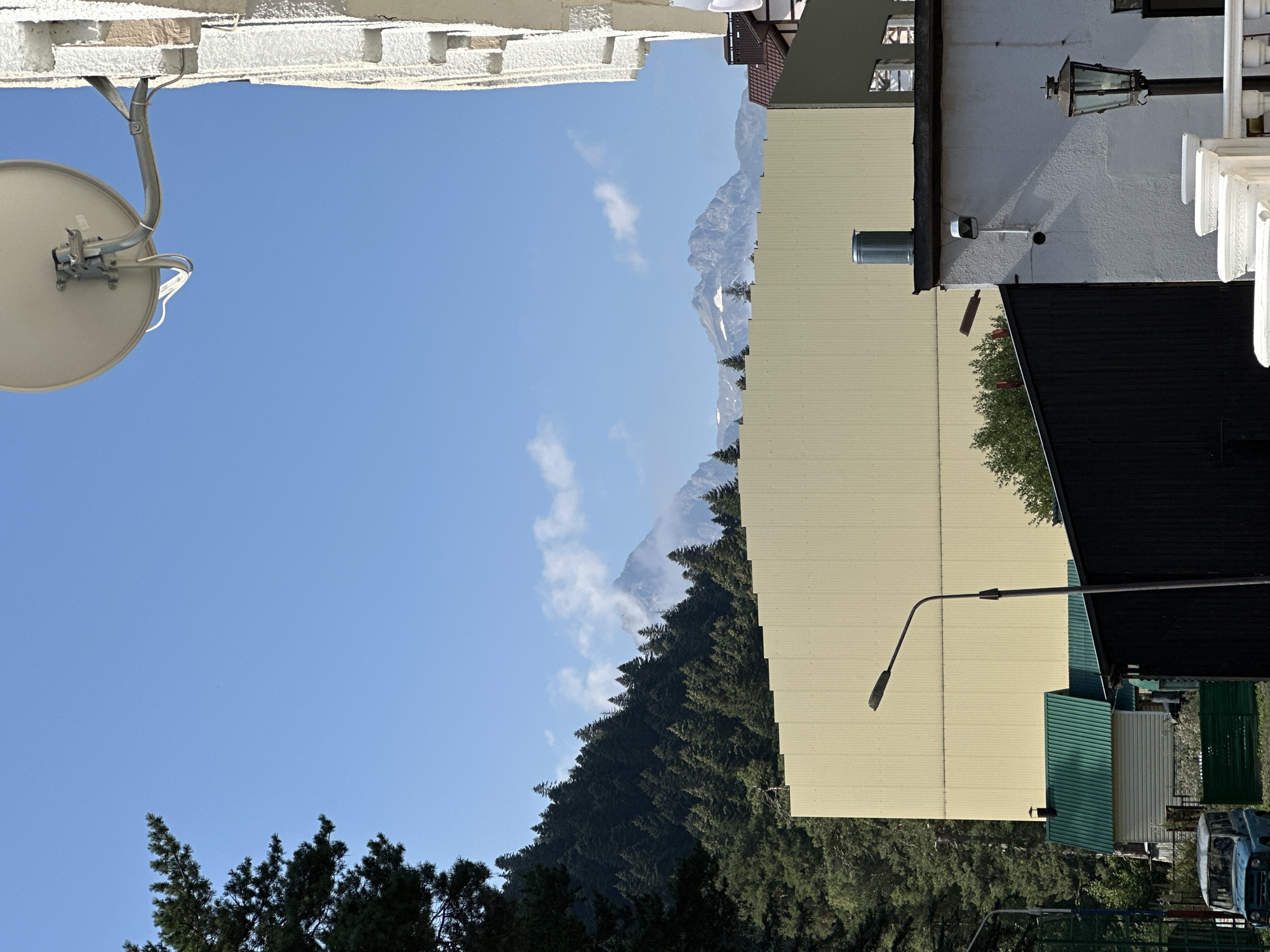 The image depicts a view of the city of Arkhyz, Russia, on a clear, sunny day. In the foreground, a portion of a white building with textured walls is visible on the right side, with a satellite dish mounted above it. A street lamp stands in the mid-ground, to the left of a black, shed-like structure. Behind these, a large, light yellow, corrugated metal wall dominates the center of the frame. To the left of this wall, a dense forest covers a hillside that slopes upwards towards a mountain range in the background. The mountains are partially obscured by white clouds and snow caps. Further to the left, a blue vehicle is parked in front of a small building with a green roof and white siding. The sky is a bright, clear blue.