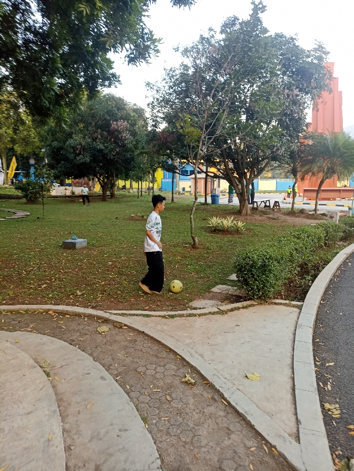 The image shows a young boy in a white shirt and black pants playing with a yellow soccer ball in a grassy park in Jatinangor, Indonesia. The boy is standing on a concrete path. The path leads to a larger grassy area, which is surrounded by trees. There are some other people in the distance. The park is near a paved road. The scene is peaceful and relaxed. The time of day is afternoon. The weather is sunny. The coordinates provided are -6.931, 107.776.