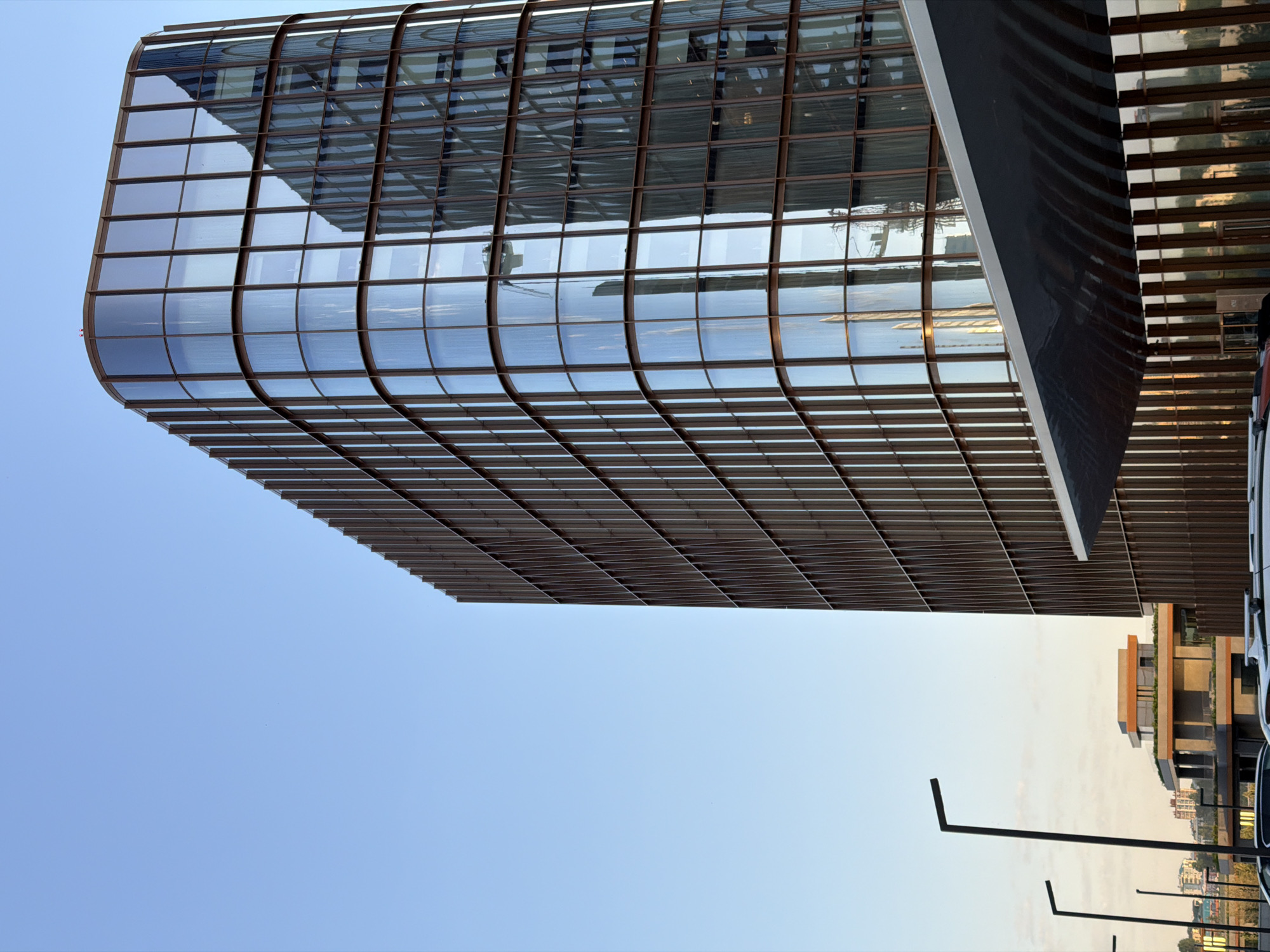 The image shows a modern building with a curved, glass facade in Kazan, Russia. The facade is segmented into a grid of glass panes with a brown frame. The windows reflect the sky and other buildings. Below the building, there are street lights and parked cars. The sky is clear and blue.