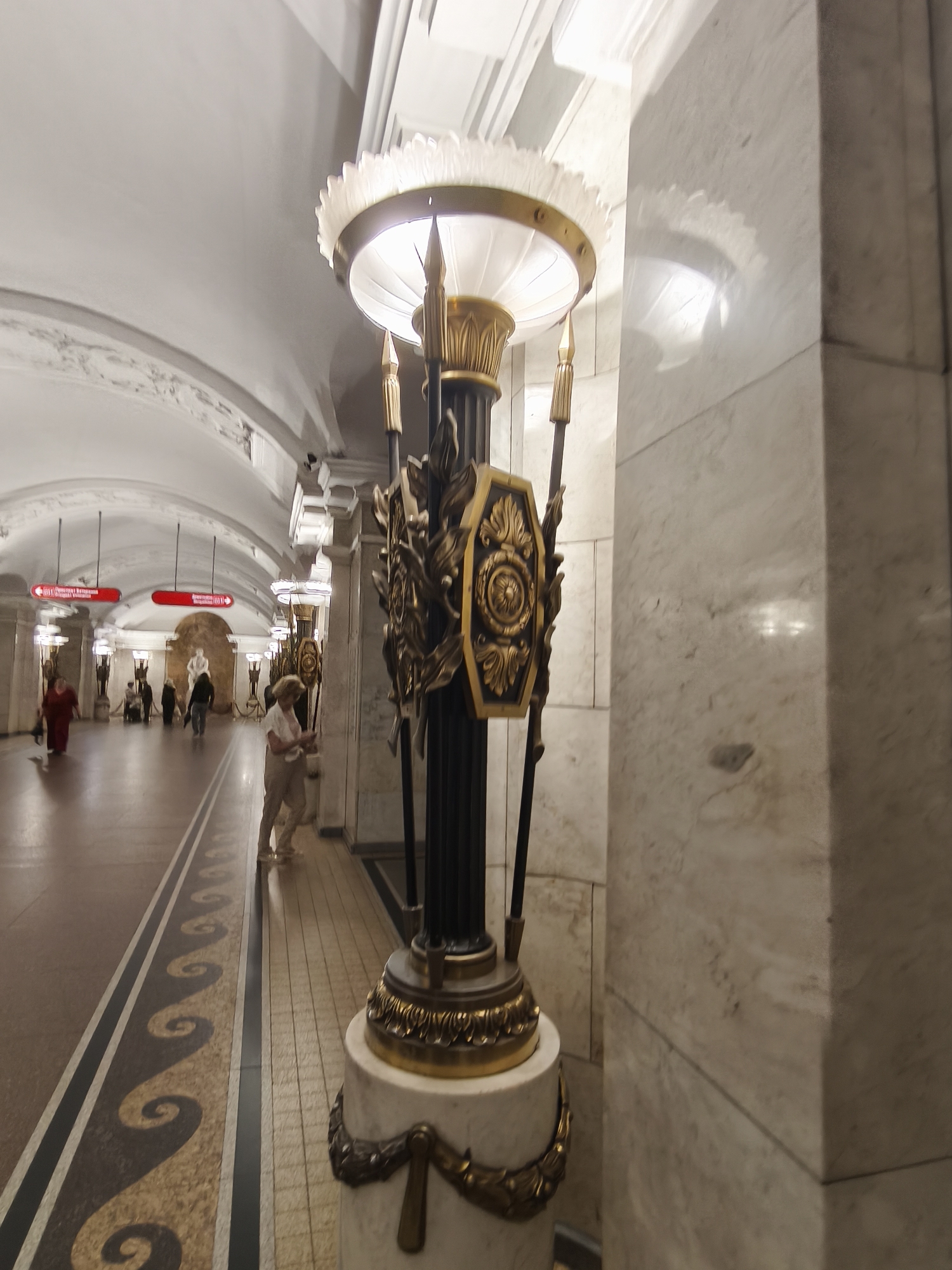 The image shows a section of a metro station in Saint Petersburg, Russia, likely on the ground floor or a mezzanine, given the architectural style. The station features high, arched ceilings with decorative molding, and the walls are clad in marble. A grand, ornate lamppost stands prominently in the foreground, made of dark metal with gold accents and featuring a large, fluted glass shade. The lamppost is positioned on a marble pedestal decorated with gold foliage.
The floor of the station is a patterned mosaic with a distinct wavy design along the walkway, separated by dark lines. In the background, several people are walking along the platform. One woman in a red dress is walking towards the camera, while others are further down the station, some appearing to be standing or walking away. Red directional signs with white lettering are visible on the ceiling. The overall impression is one of grandeur and historical significance, characteristic of many Saint Petersburg metro stations.
