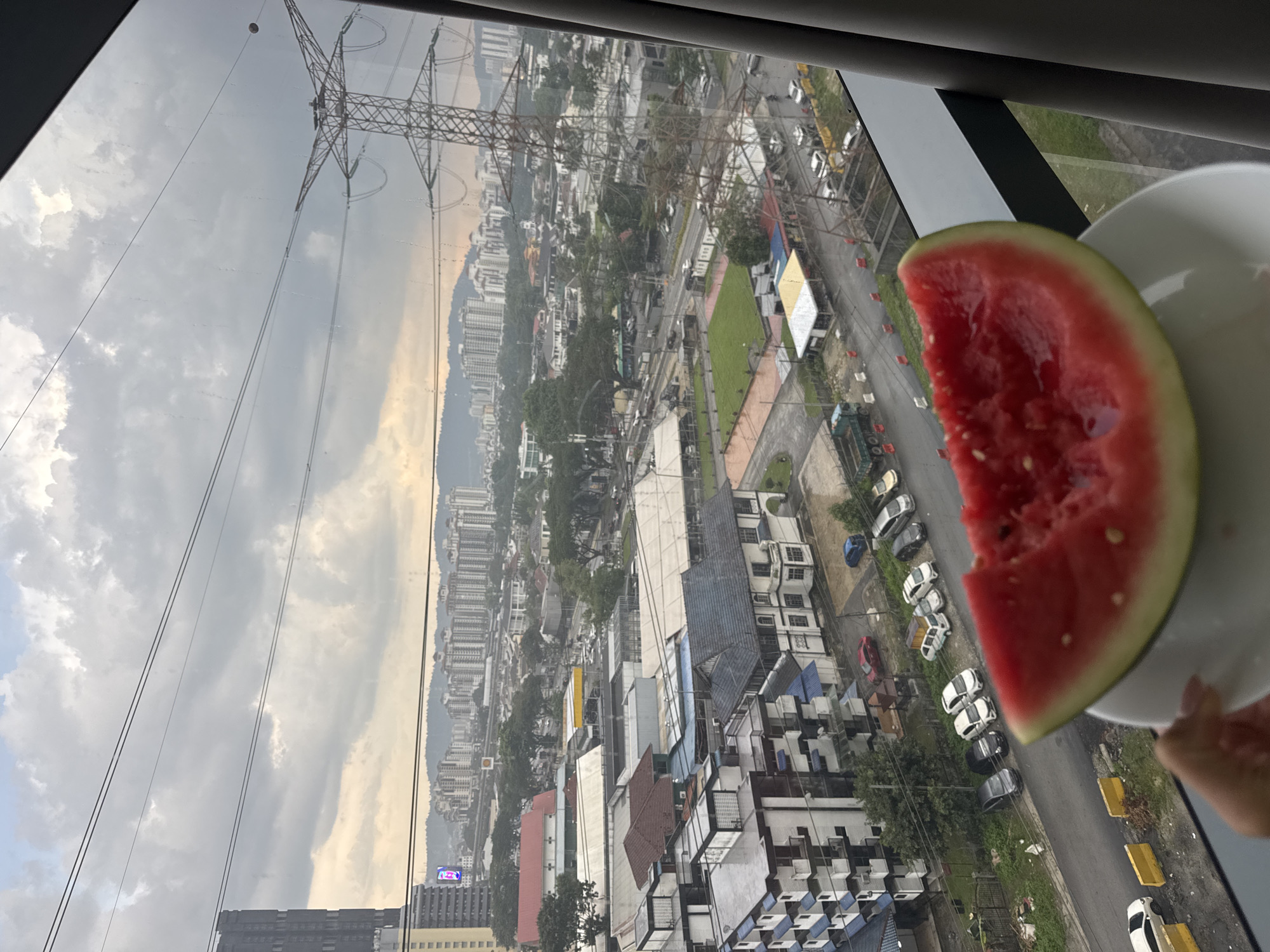 The image, oriented on its side, presents an expansive urban landscape viewed from a high vantage point, likely a window, with its frame visible at the edges. When rotated clockwise to an upright position, the scene reveals a cloudy, overcast sky dominating the upper portion. In the bottom right foreground, a hand holds a white plate with a partially eaten slice of red watermelon.
The sprawling cityscape, consistent with Kuala Lumpur, Malaysia, stretches below. A prominent electrical transmission tower with multiple power lines cuts across the left and center, extending into the distant urban environment. The city consists of a dense mix of residential buildings, including apartment blocks and smaller houses with varied roof colors, interspersed with commercial structures and patches of green landscaping. Numerous cars are parked along the visible streets. A faint "SHELL" logo is discernible on a red-roofed building in the midground. In the hazy distance, a range of hills or mountains forms the horizon. The scene depicts a daytime view under an overcast sky, capturing a moment of enjoying a snack against the backdrop of a vast city.