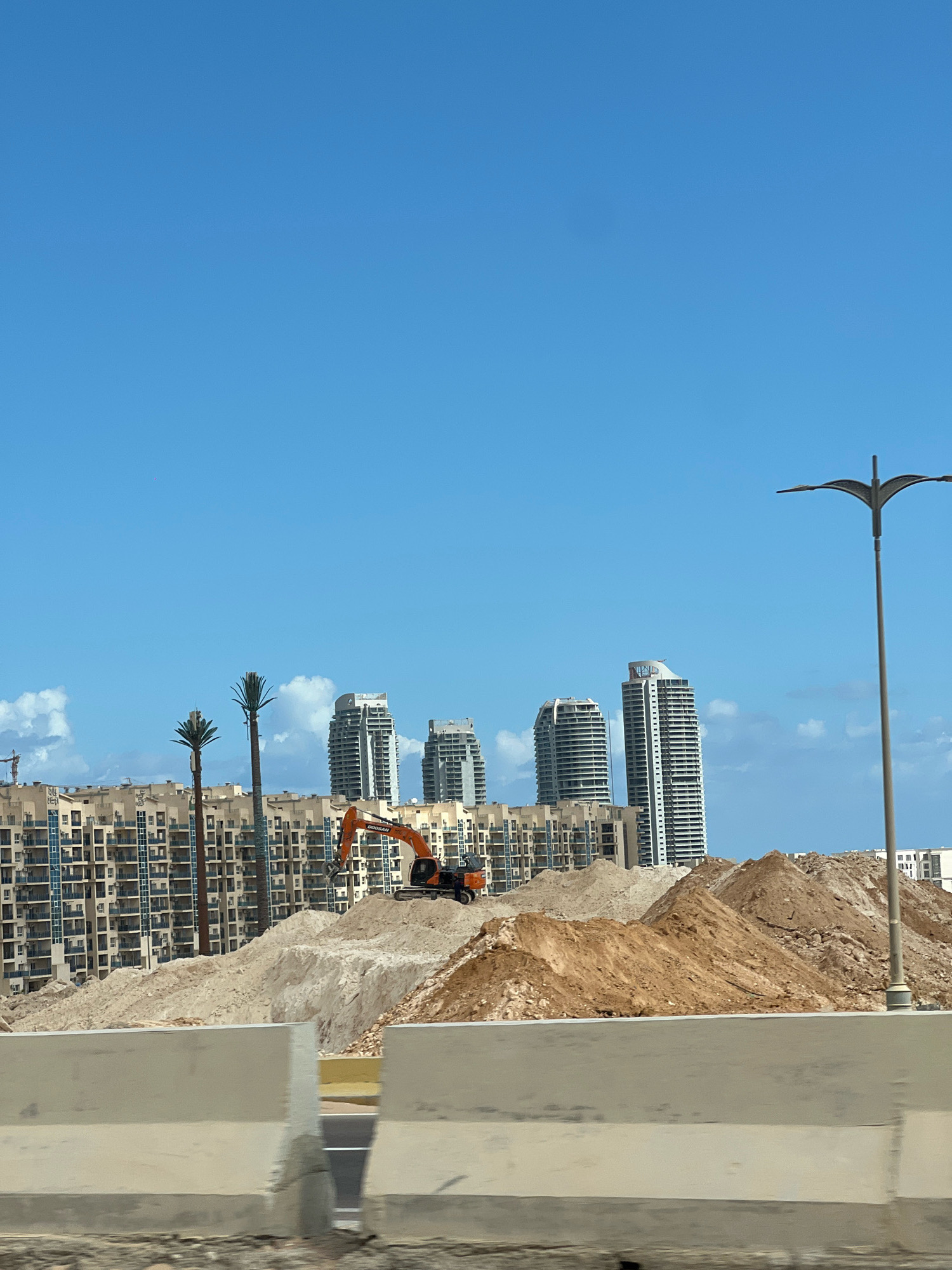 The image depicts a construction site in Sidi Abdel Rahman, Egypt, under a clear blue sky. In the foreground, a concrete barrier runs horizontally across the frame. Behind the barrier, large mounds of earth and sand dominate the landscape, indicating ongoing excavation or land preparation.
A bright orange excavator is visible on top of one of the sand mounds, with the brand name "DOOSAN" clearly marked on its arm, suggesting active construction work.
In the midground, a large, multi-story residential building complex with a tan facade and numerous balconies is under construction or recently completed. Two tall, stylized palm trees with what appear to be communication equipment at their tops stand in front of this complex.
Further in the background, several modern high-rise buildings with white and grey exteriors rise against the sky, some of which appear to be hotels or luxury apartments. A construction crane is visible to the far left, partially obscured by the buildings.
A street light with a distinctive, wing-like design stands to the right of the frame, its pole extending vertically. The overall scene is one of urban development and construction, characteristic of a growing coastal area.